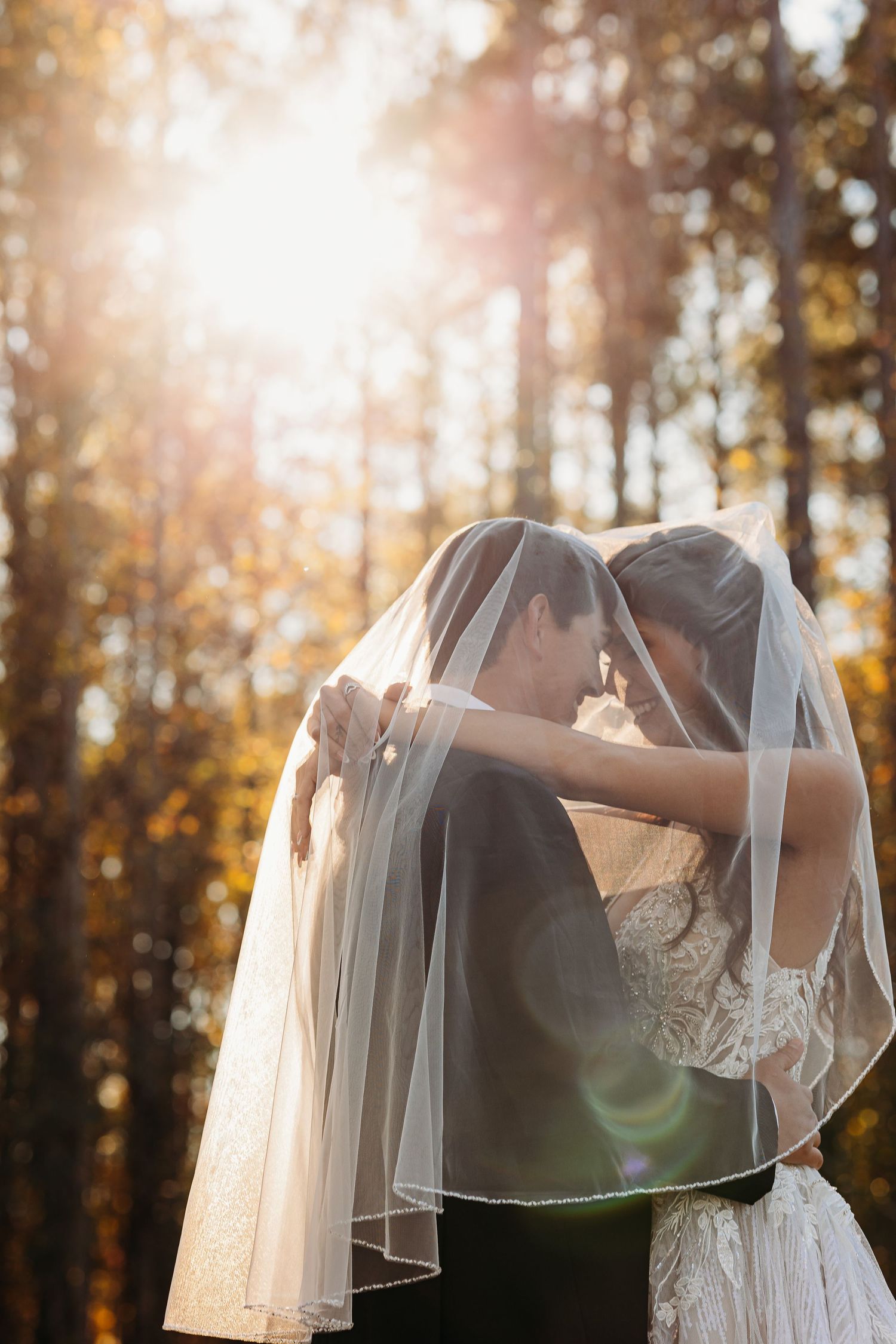 A romantic wedding couple embraces in a sunlit autumn forest with a flowing white veil creating a dreamy atmosphere.