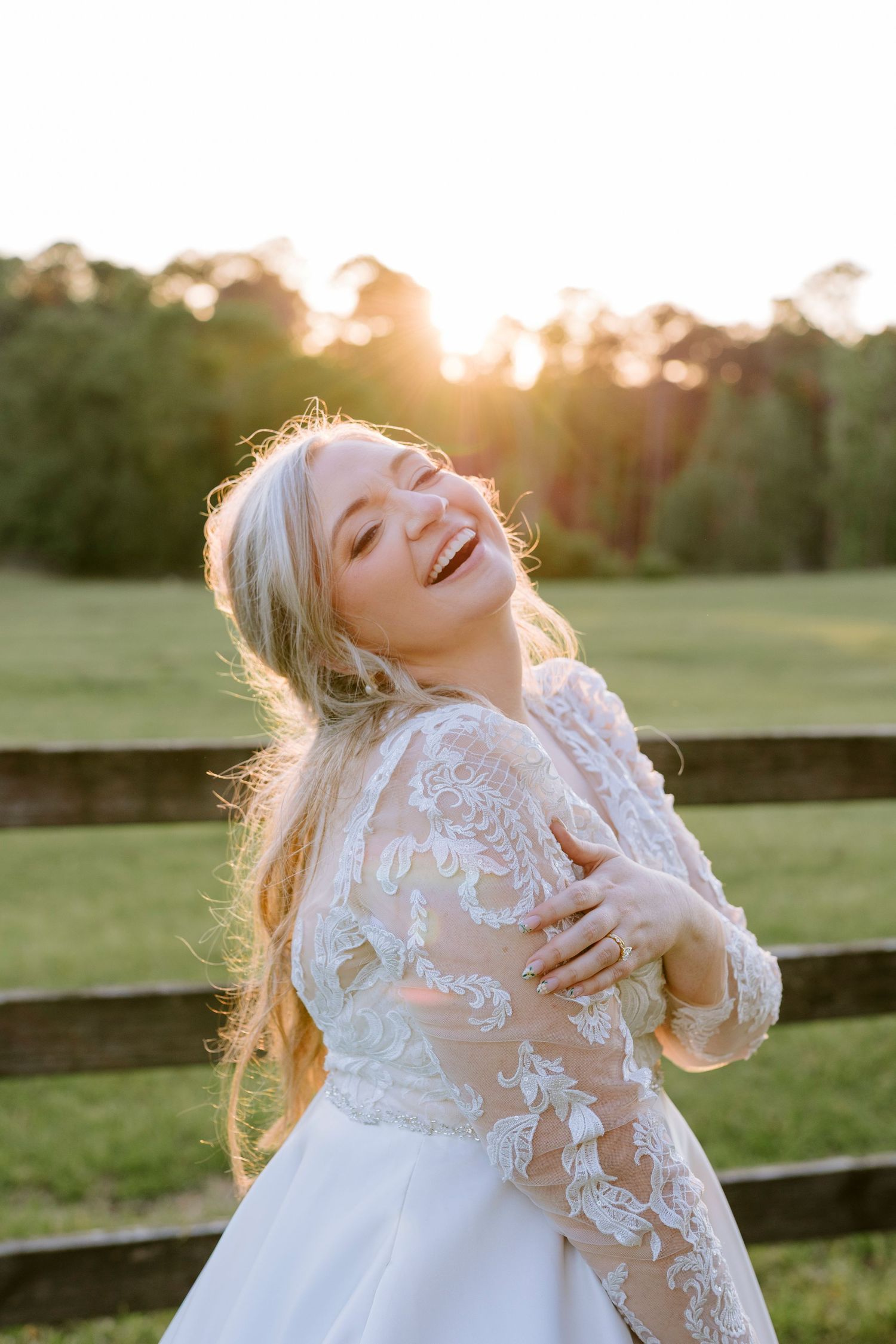 Joyful bride in lace wedding dress laughs while embracing herself in golden sunset light at outdoor venue.