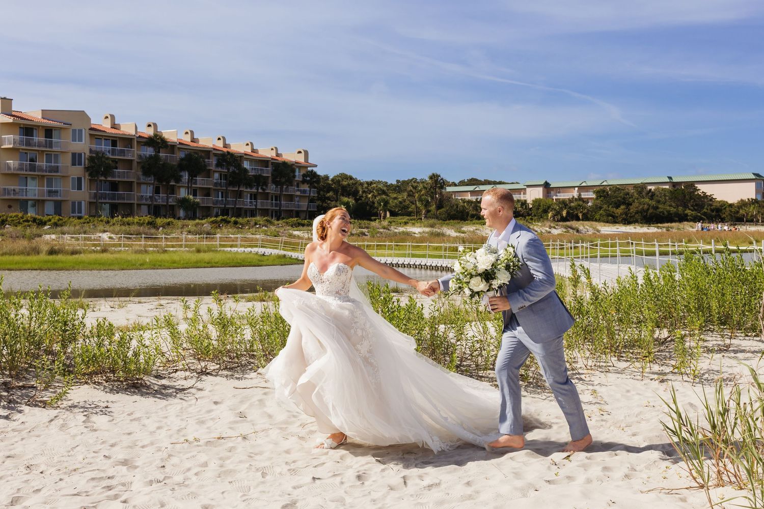 A couple in wedding attire walks along a scenic beach path with sand dunes and blue skies in the background.