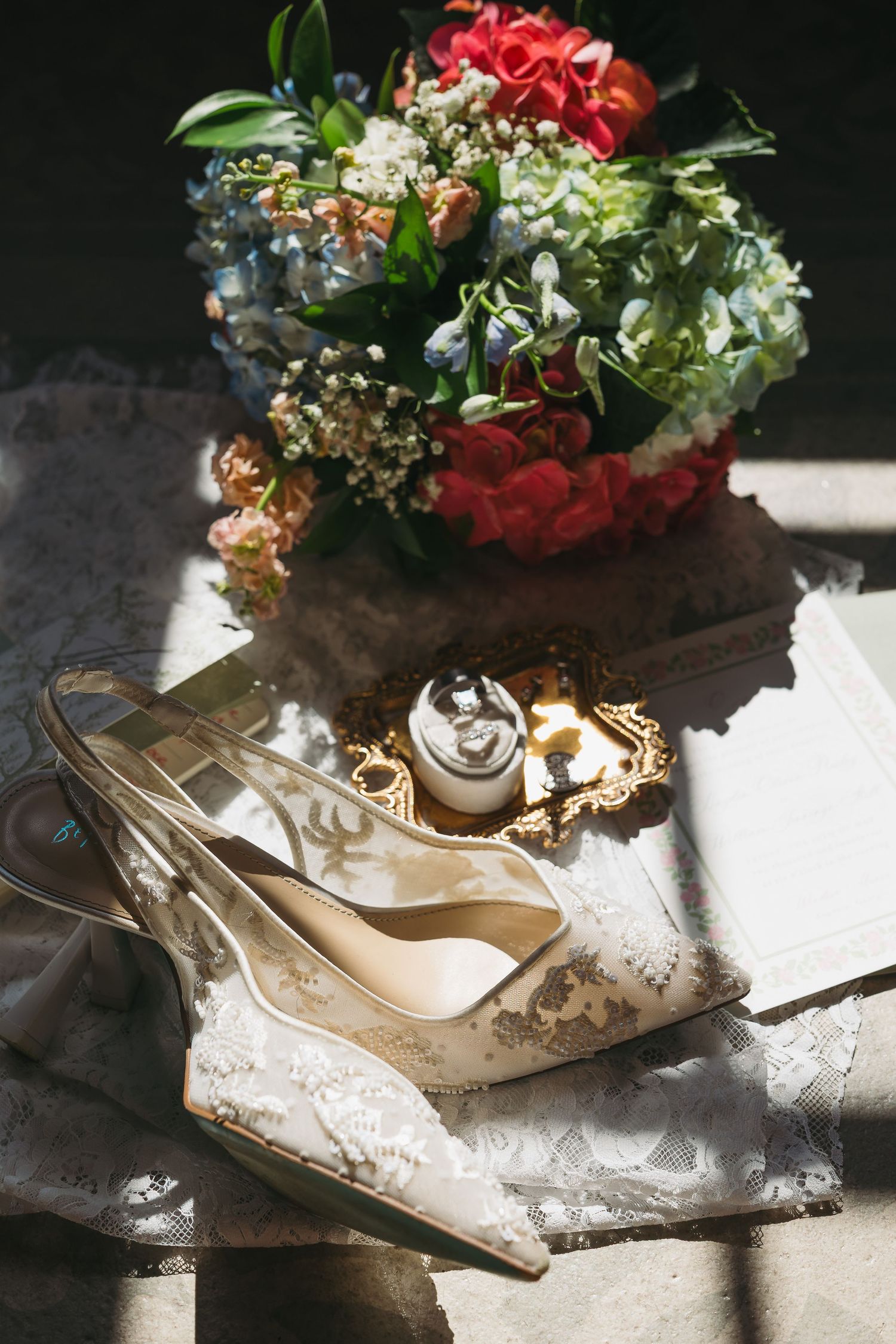 Elegant bridal flat lay with lace heels, wedding rings on gold tray, and colorful floral bouquet in natural light.