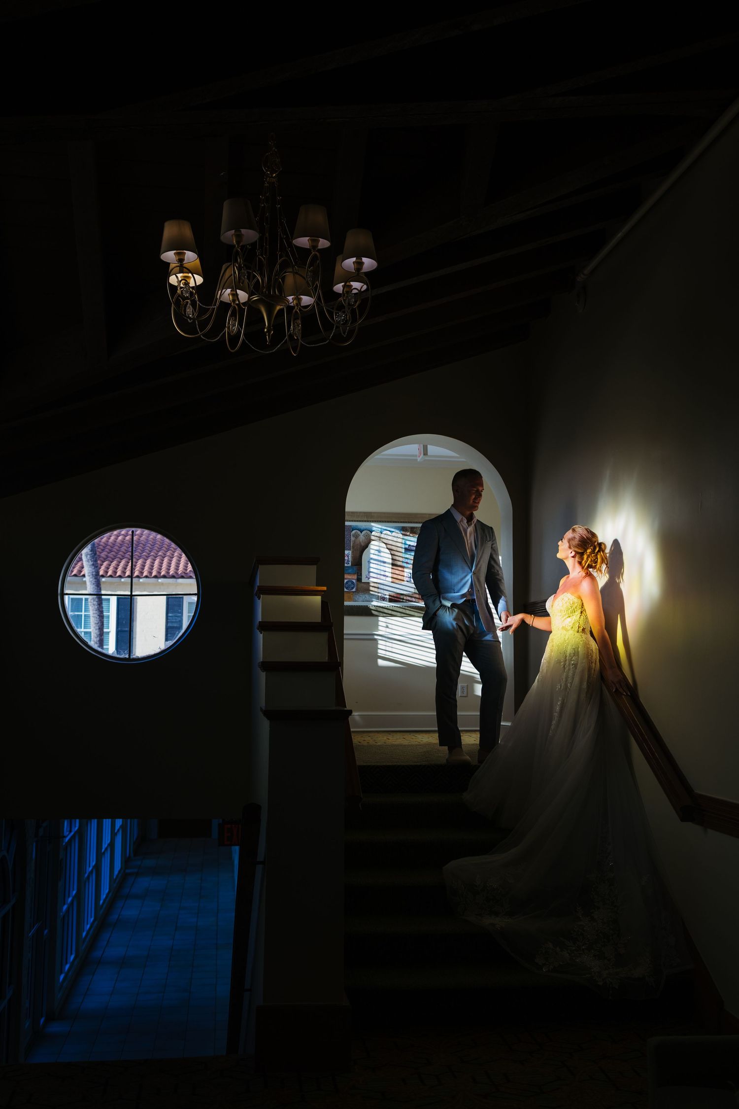 Bride and groom holding hands on a dramatic staircase with chandelier and arched window in elegant venue.