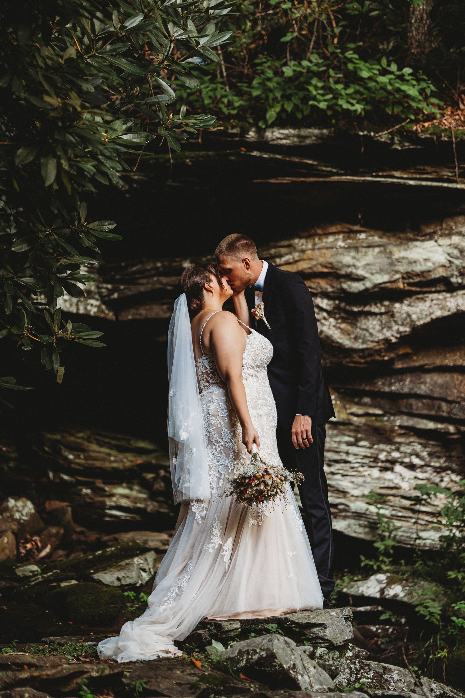 A romantic wedding couple stands intimately together against a rocky cliff wall surrounded by lush greenery.