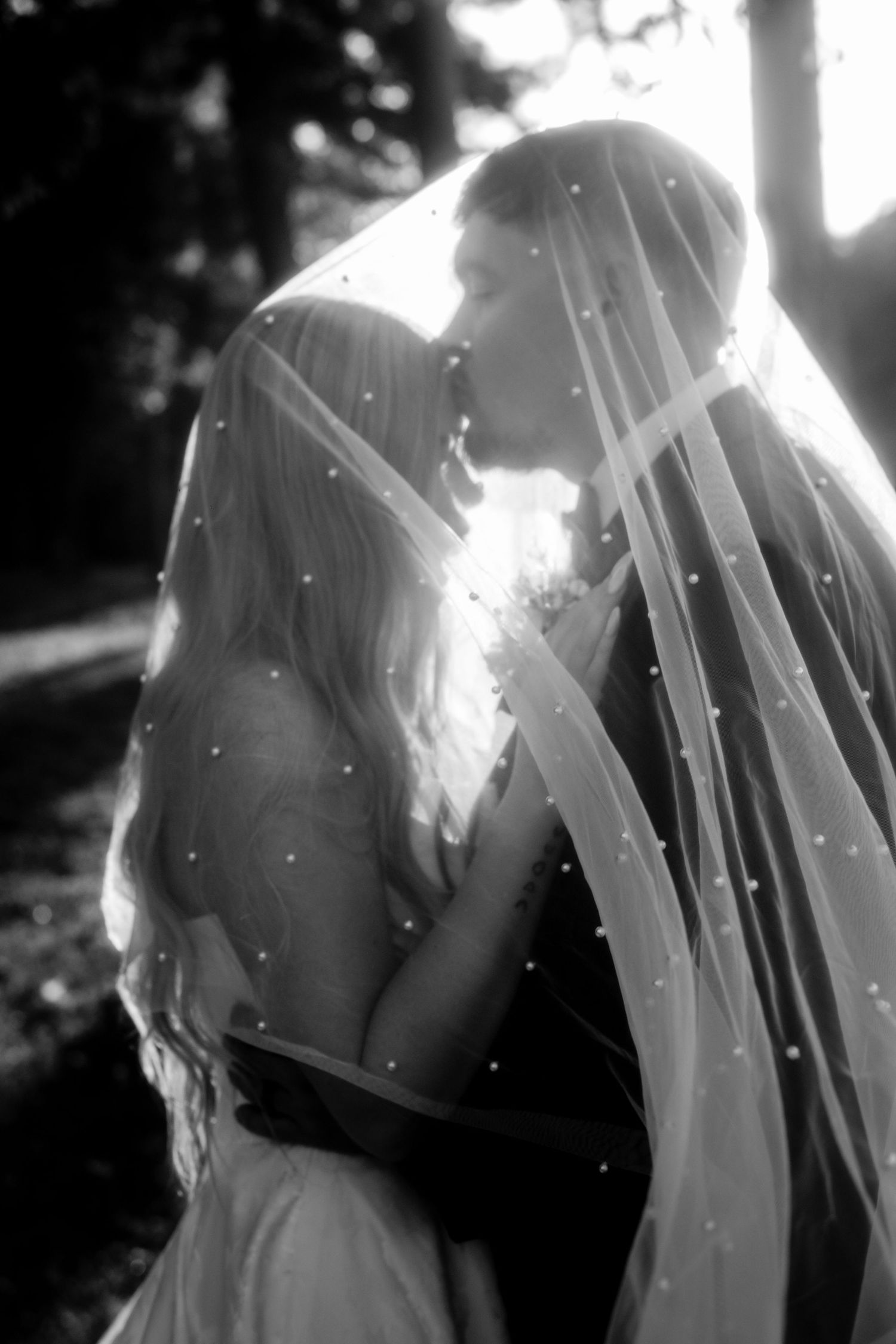Black and white photo of bride and groom sharing a kiss under a pearl-dotted wedding veil outdoors.