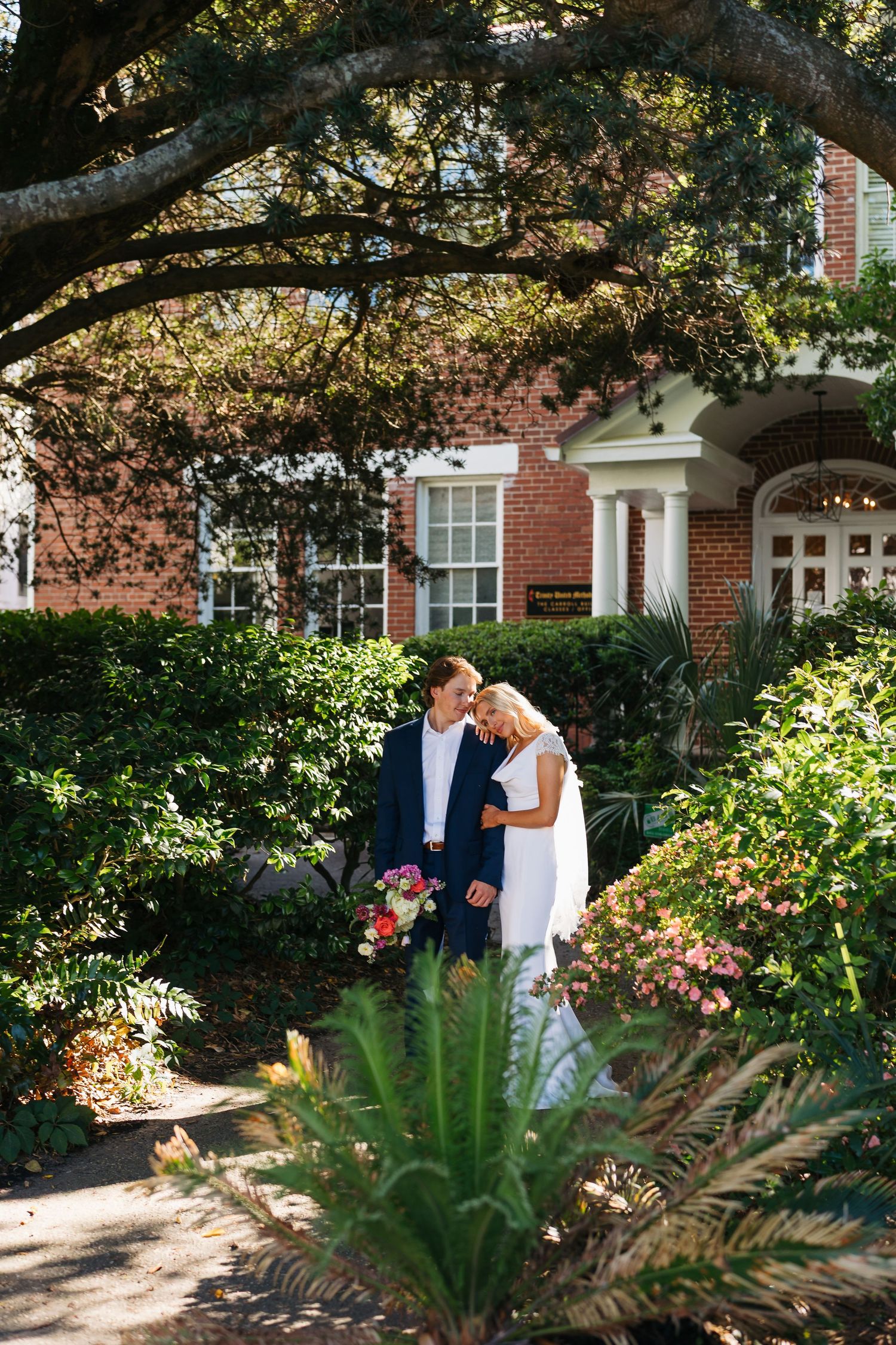 A bride and groom embrace in a lush garden outside a brick building on their wedding day.