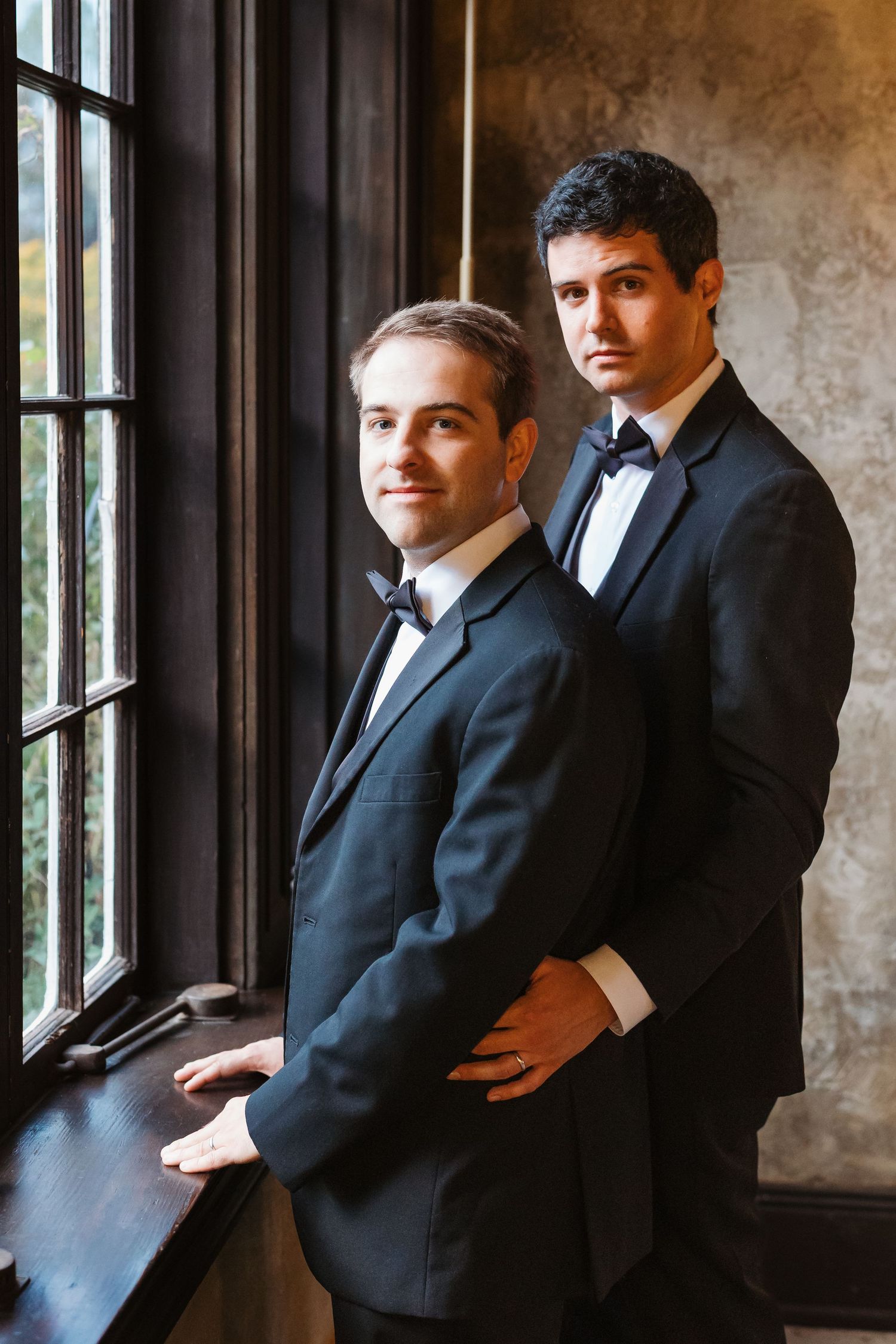 Two men in black tuxedos with bow ties pose elegantly near a window in a dimly lit room.