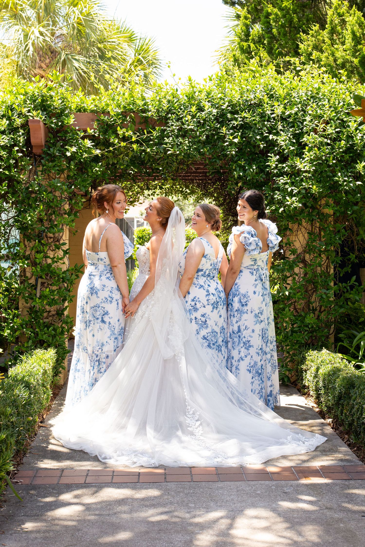 A group in blue floral bridesmaid dresses surrounds a bride in white on a sunlit garden path.