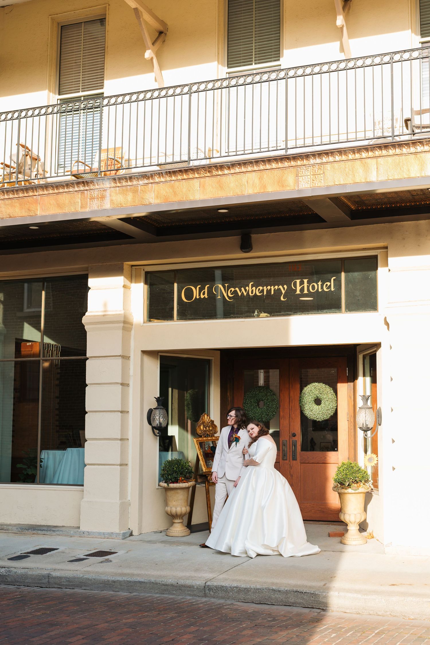 Two brides pose in wedding attire outside the historic Old Newberry Hotel entrance with elegant decor.