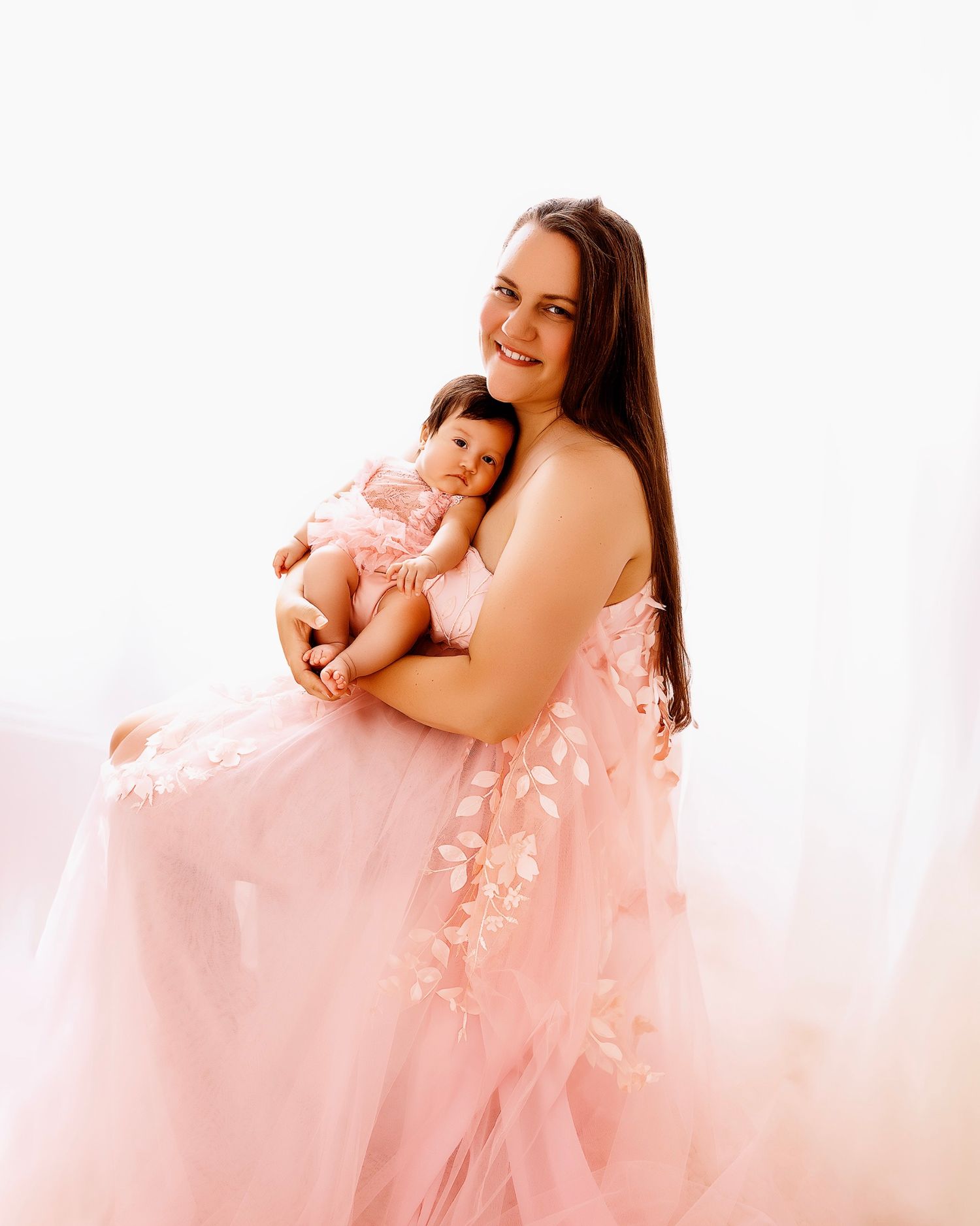 Parent and infant in pink floral dress pose against light background.