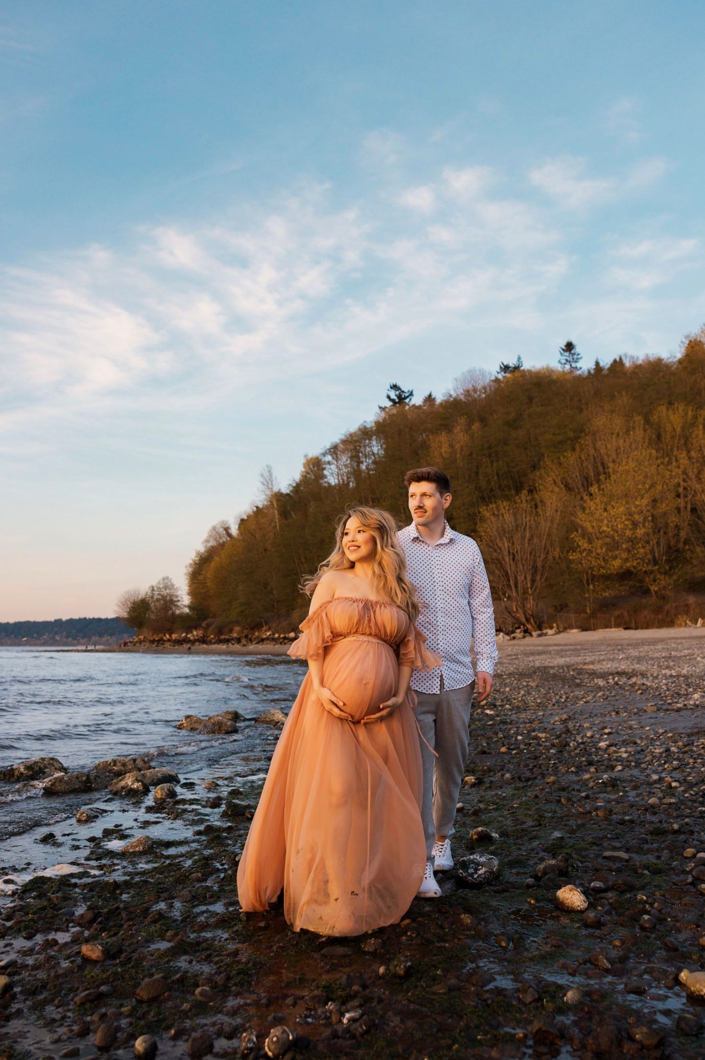 Pregnant couple walking on a rocky beach at sunset, woman in flowing peach maternity gown cradling her baby bump.