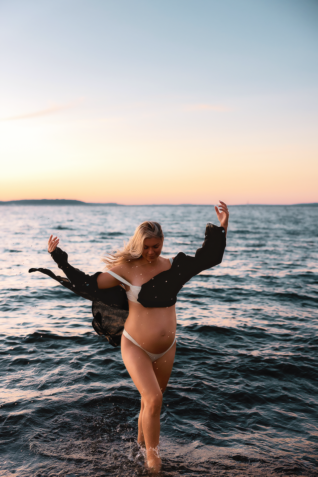 Pregnant woman in bikini and open black shirt dancing joyfully in ocean waves at sunset.