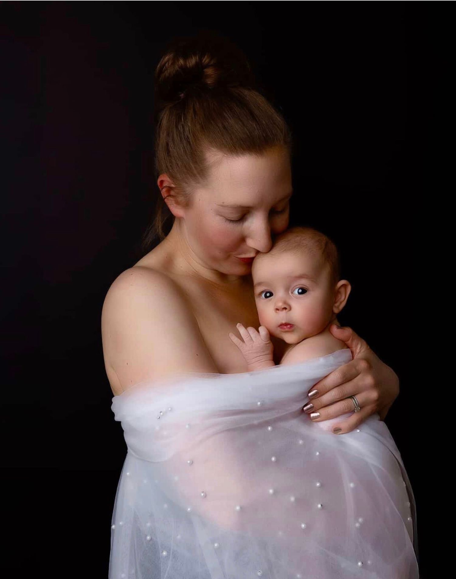 A mother tenderly kisses her baby wrapped in a white pearl-adorned fabric against a dark background.