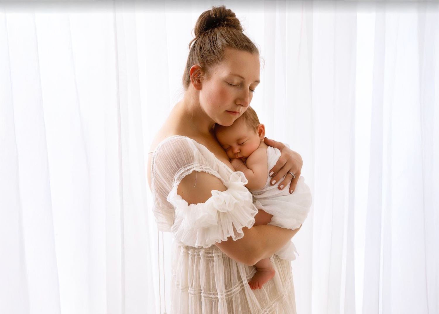A mother in a white dress tenderly holds her sleeping newborn baby against her chest near sheer curtains.