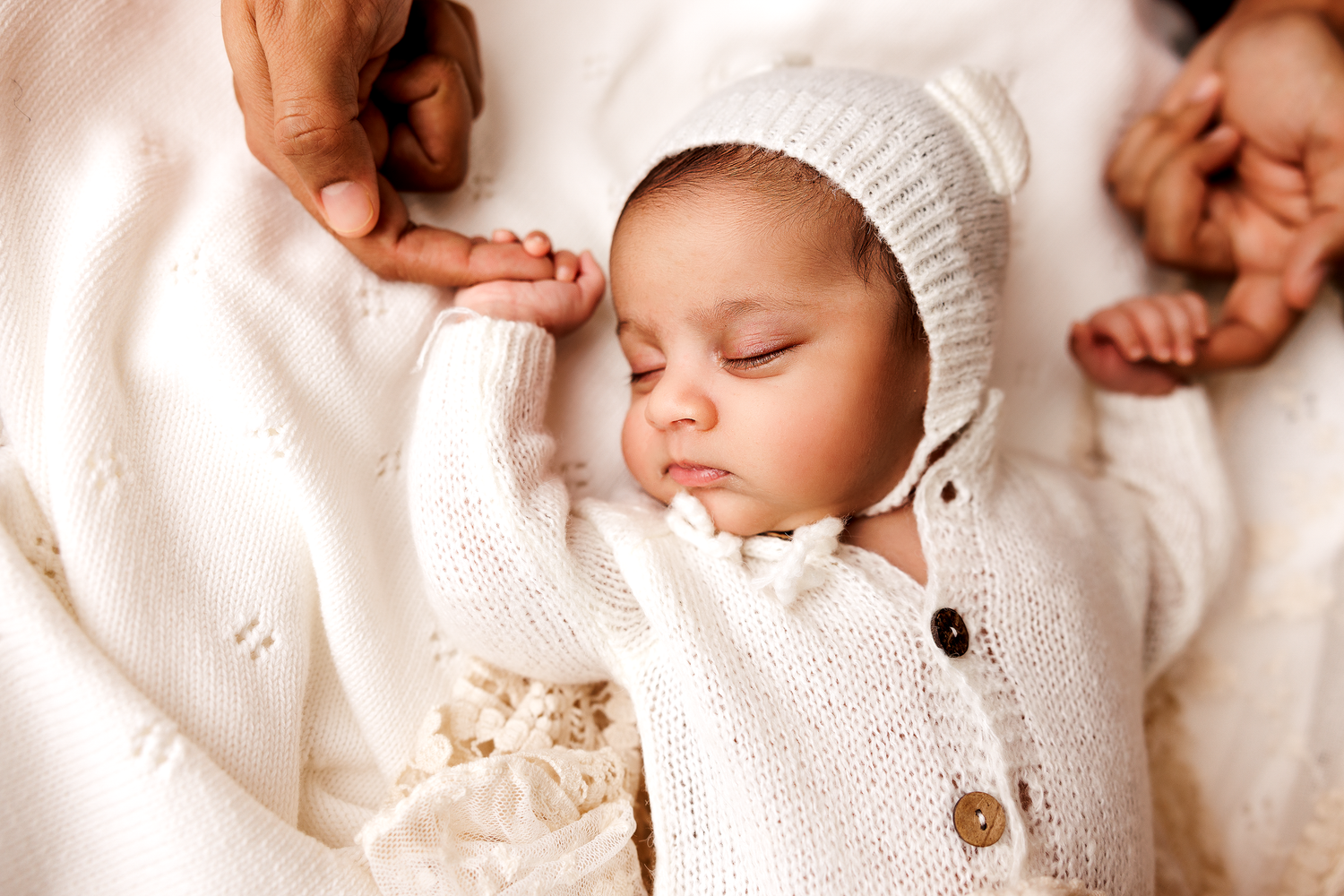 Sleeping newborn baby in white knit outfit and bonnet held by parents hands on soft white blanket.