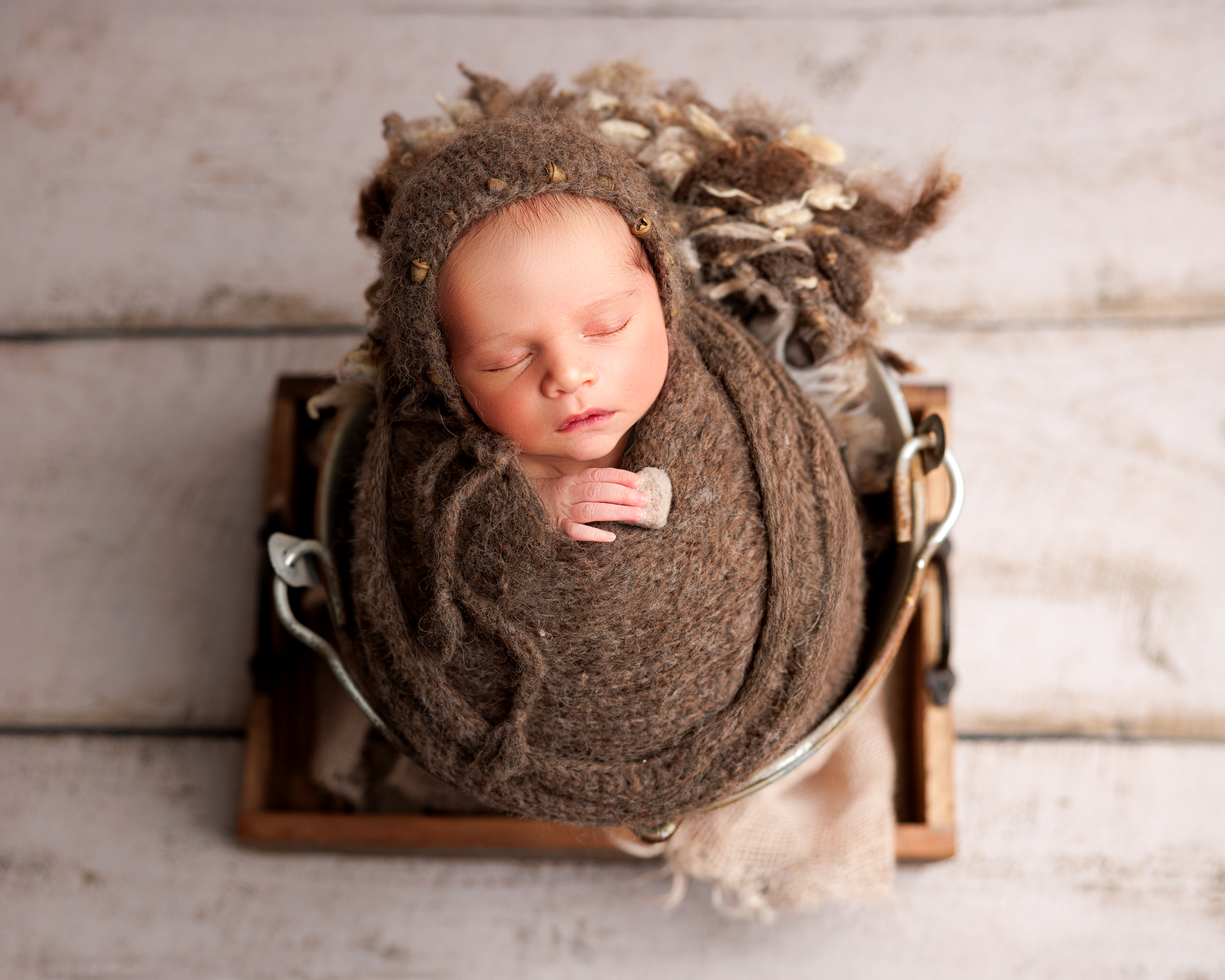 Sleeping baby wrapped in soft brown knit wrap surrounded on wooden backdrop.
