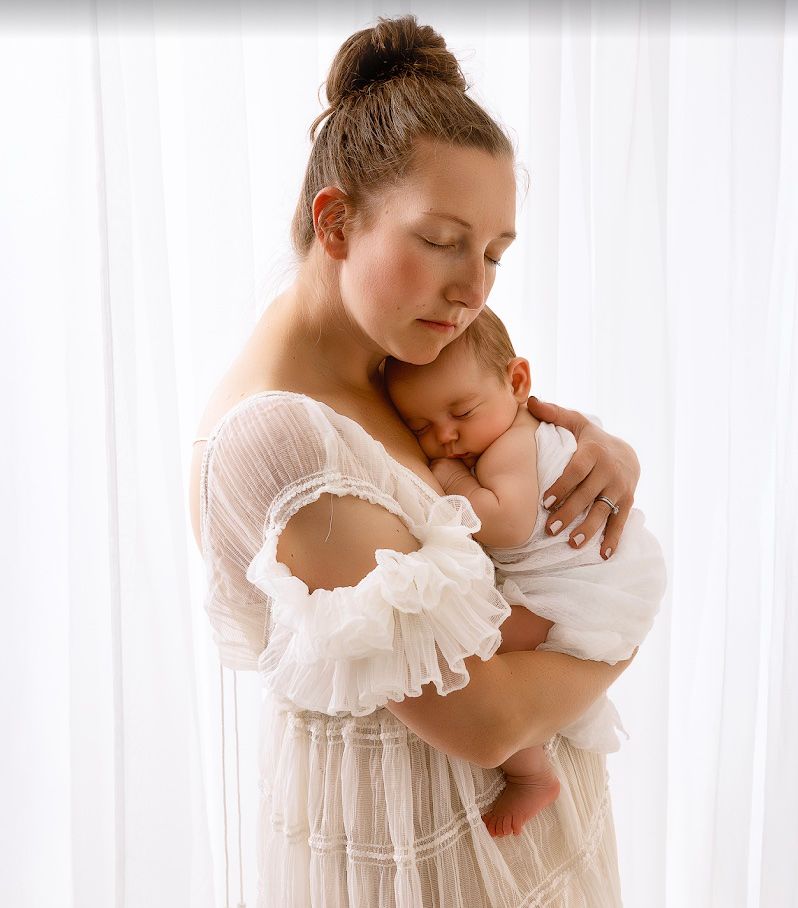 A tender moment captured in soft light as a parent in white embraces a sleeping newborn against a bright backdrop.