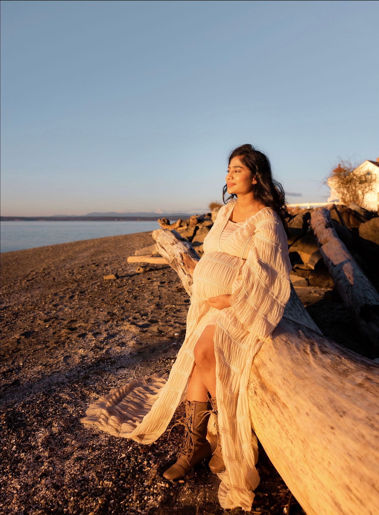 Pregnant woman in white lace dress and boots sits on driftwood at golden hour beach, gazing at the horizon.