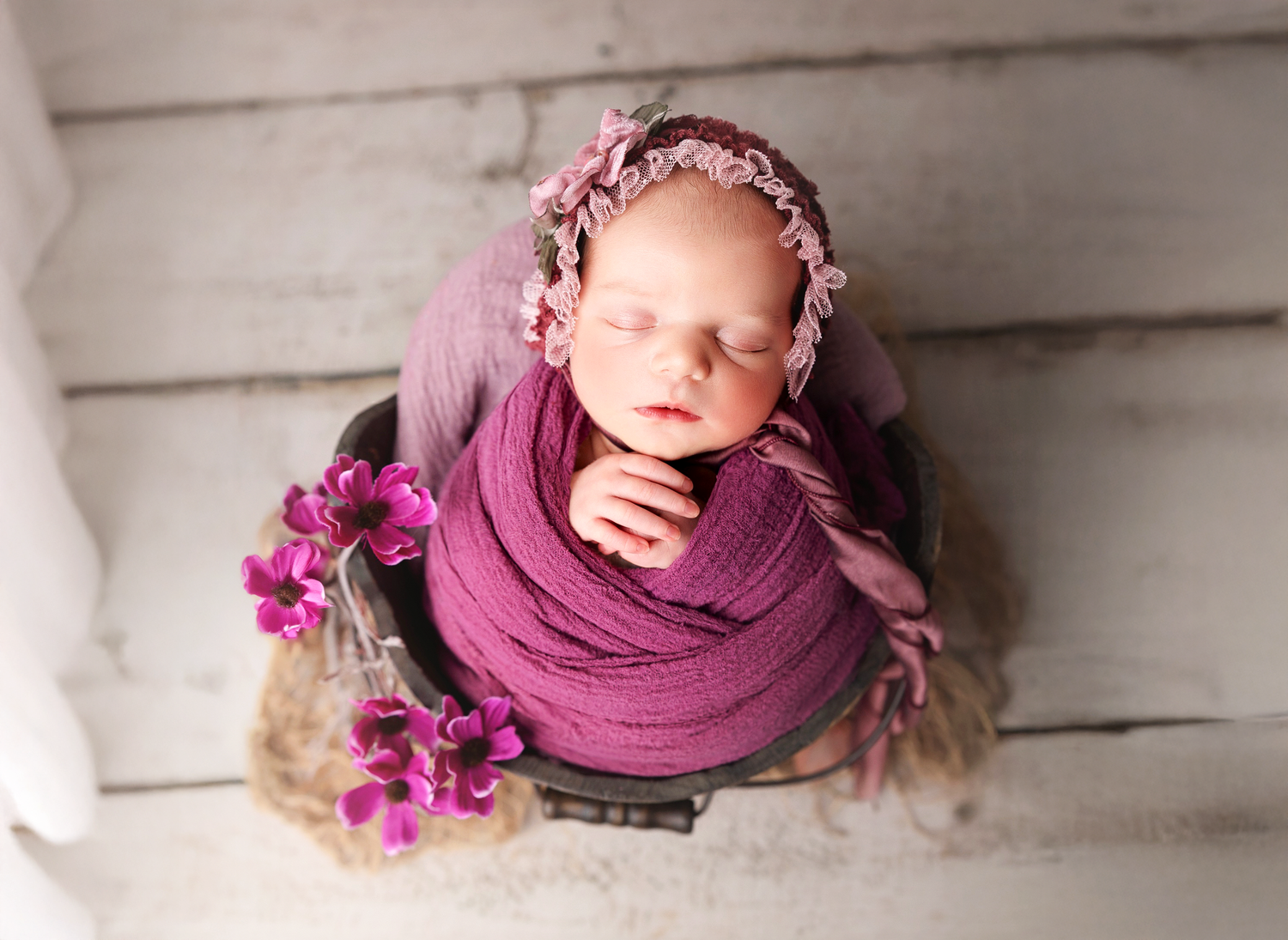 Newborn baby girl swaddled in purple wrap with floral headband, sleeping peacefully in a rustic bucket.
