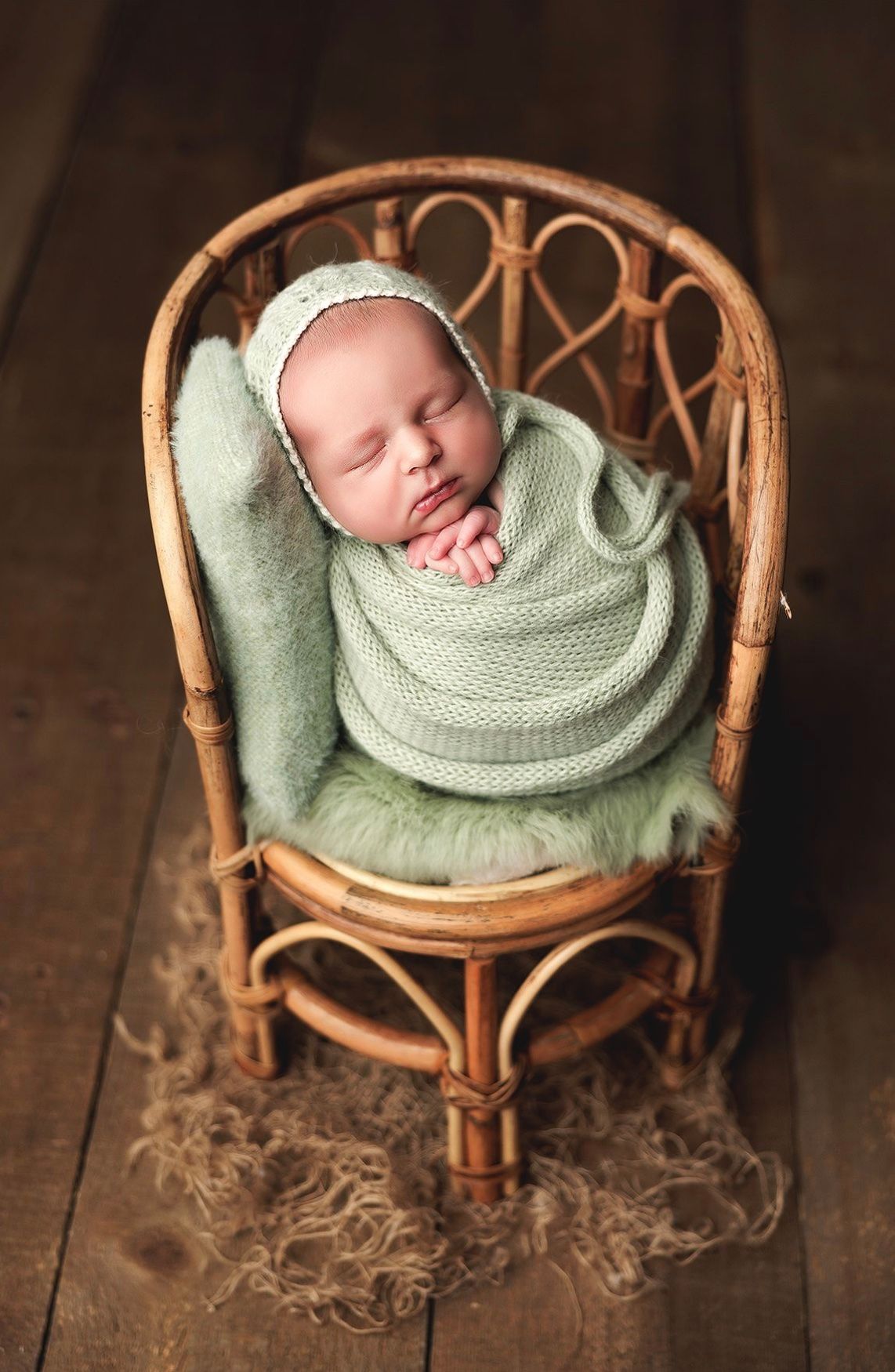Newborn baby swaddled in mint green wrap and bonnet, sleeping peacefully in a small rattan chair on wood floor.