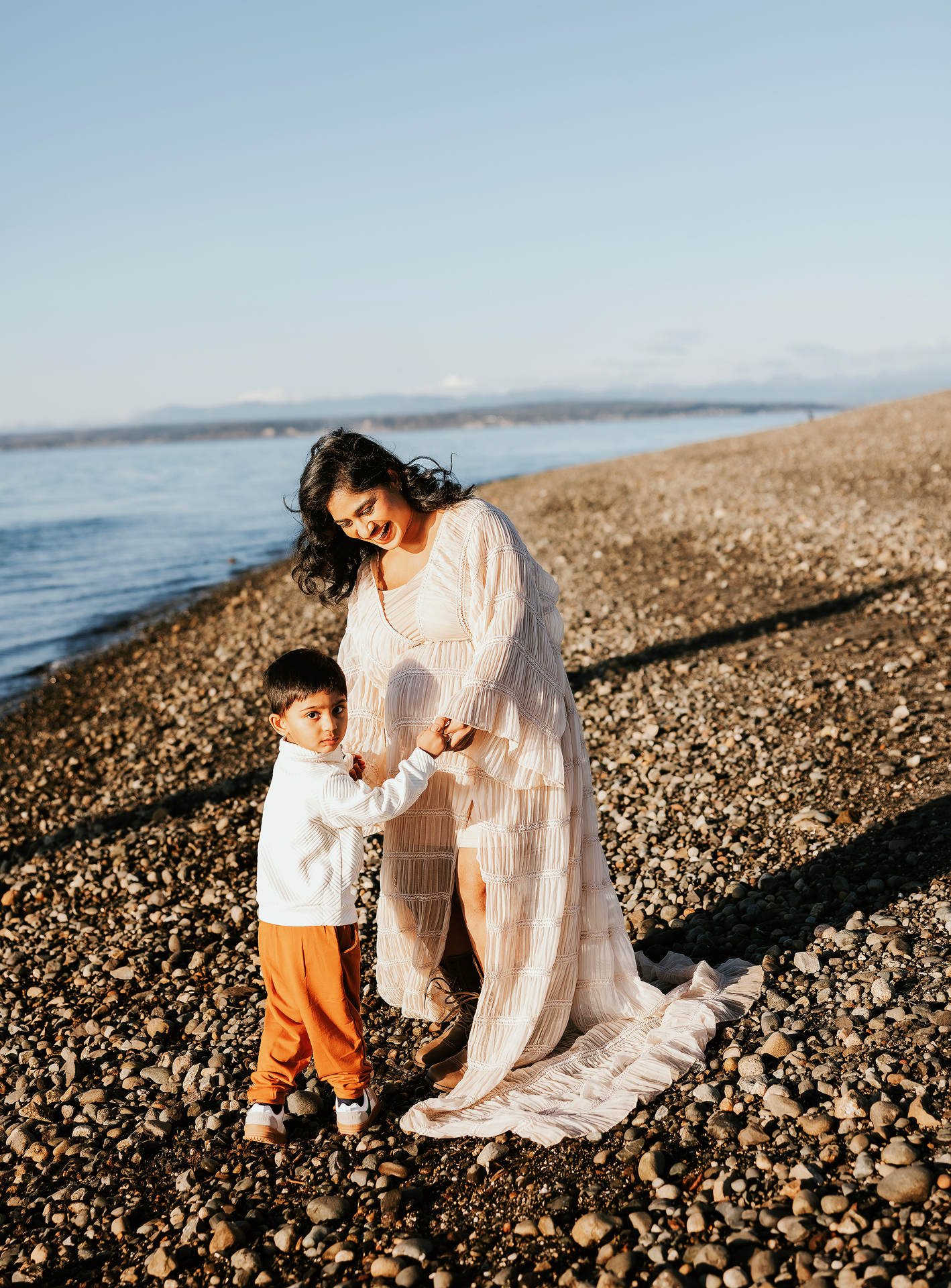 A mother in a white dress holds hands with her toddler in orange pants on a rocky beach shoreline.