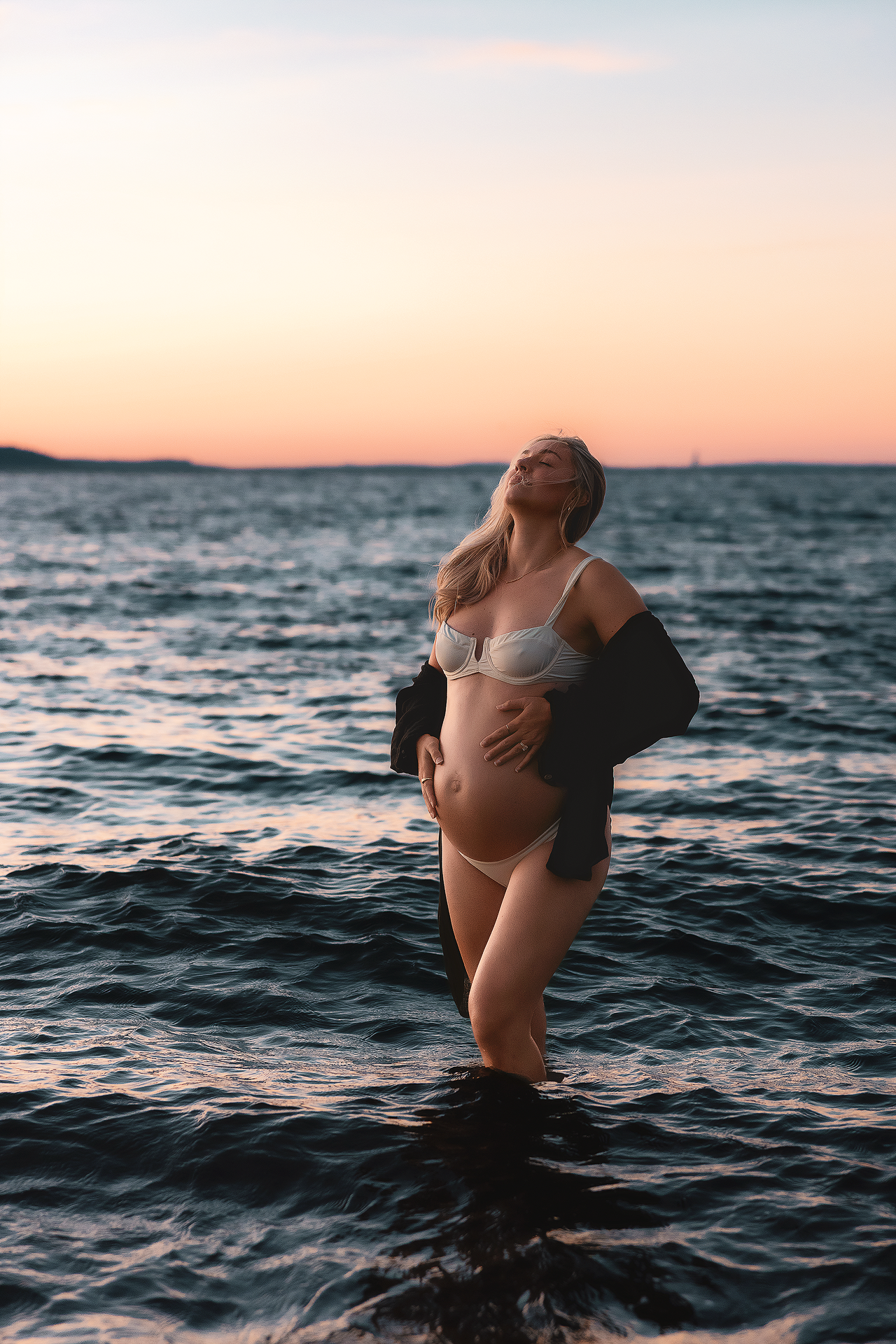 Pregnant woman in white lingerie standing in ocean waves at sunset, cradling her baby bump with flowing blonde hair.