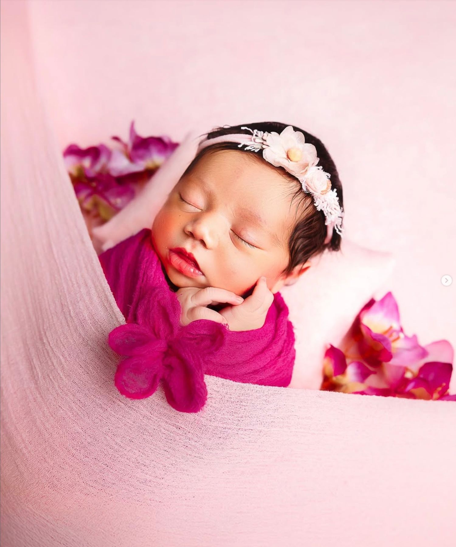 A sleeping newborn in a pink and magenta setting with floral accents and delicate headband.