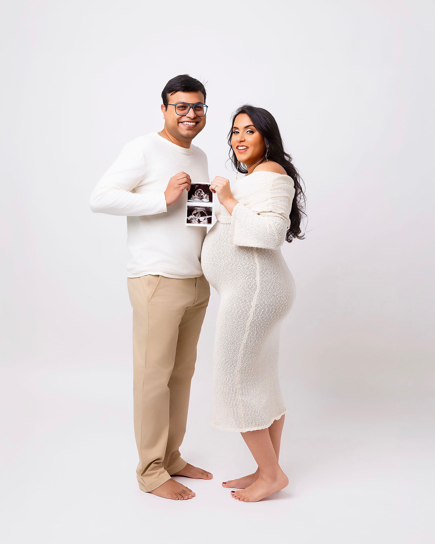 A couple in white outfits poses together for pregnancy announcement photos against a light background.