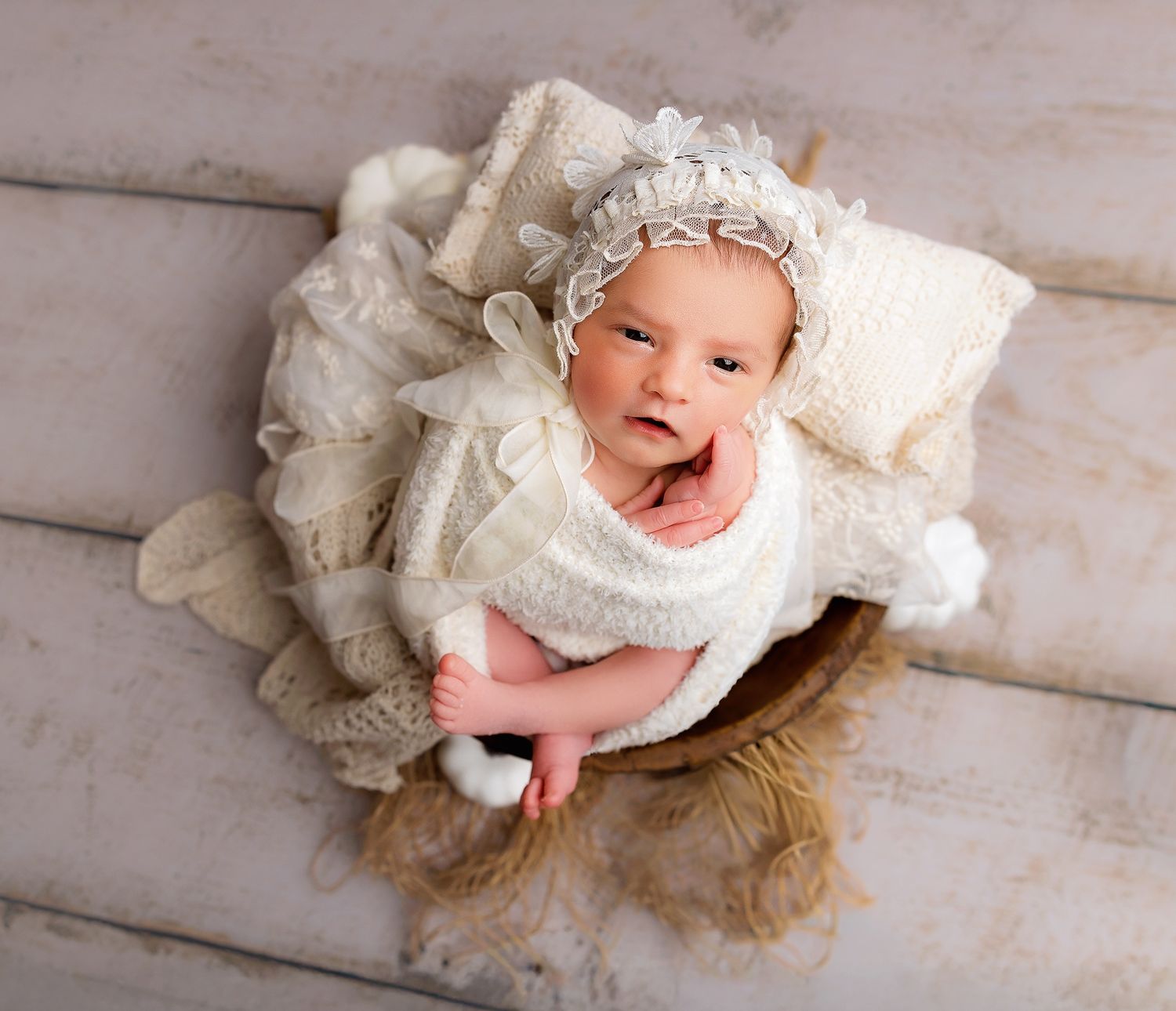 A baby wearing a white lace bonnet and dress lies in a wooden basket with cream-colored fabric on a light wooden floor.