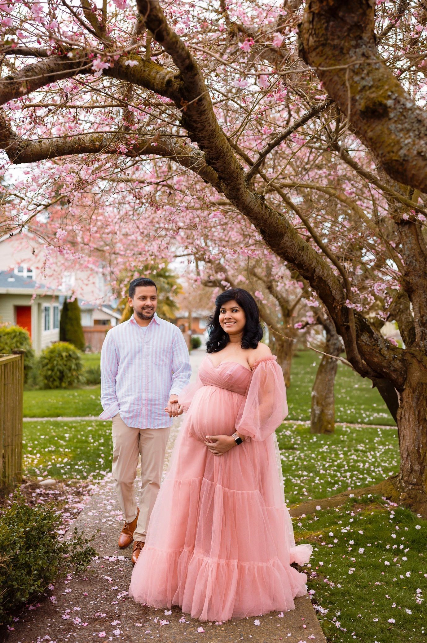 A couple poses under blooming pink cherry blossom trees in a spring garden with scattered white flowers on the ground.