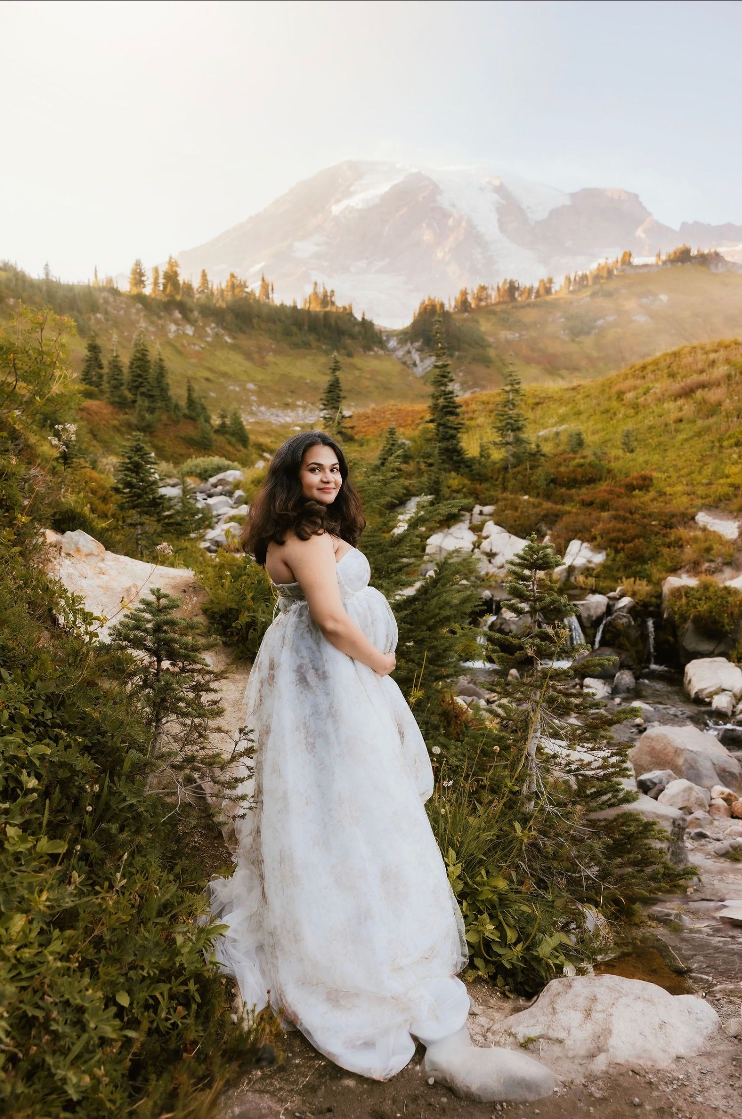 A person in a flowing white wedding dress poses among mountain scenery with evergreen trees and rocky terrain at sunset.