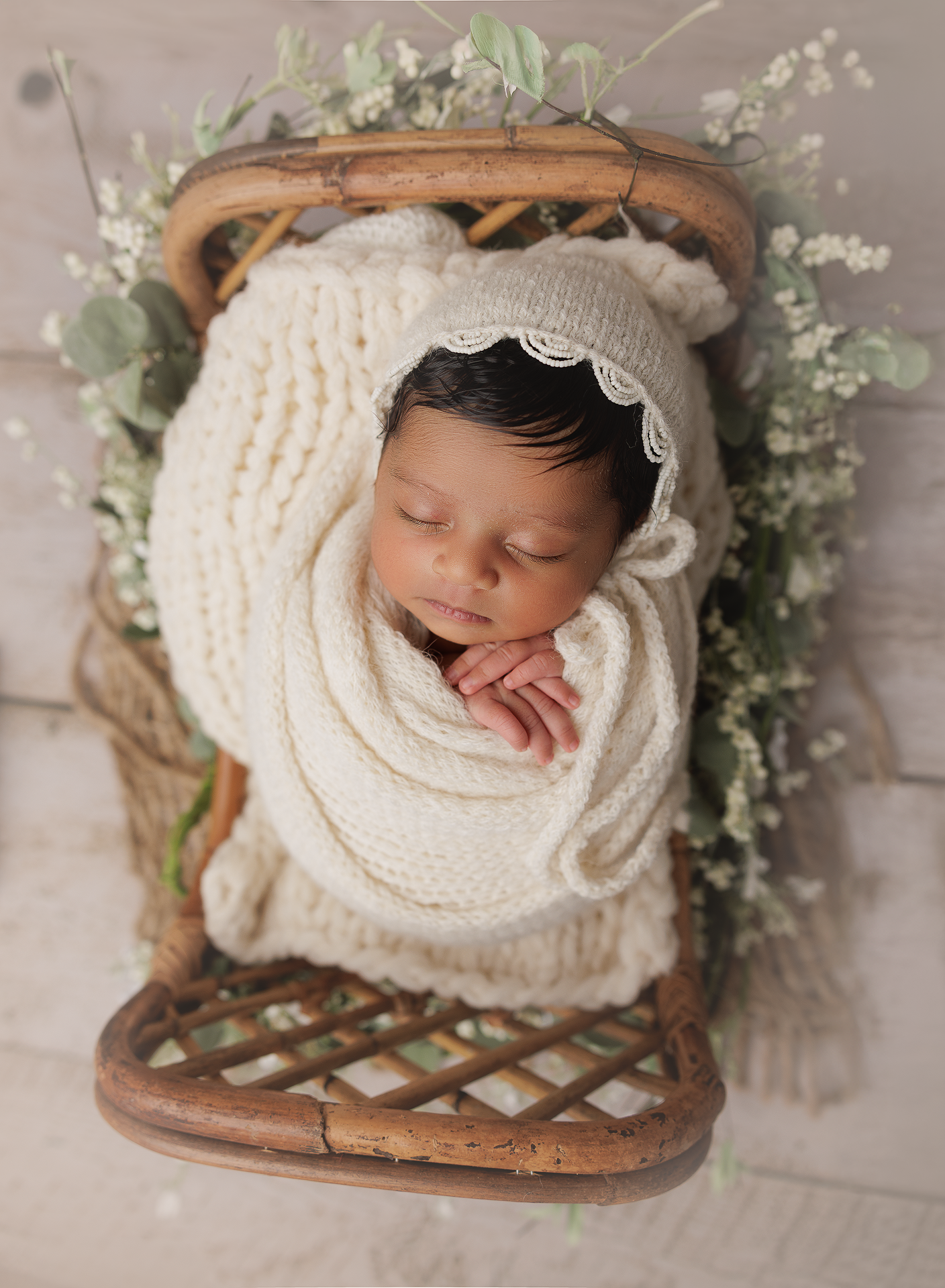 Newborn baby sleeping peacefully wrapped in cream knit blanket with delicate white flowers in wooden basket setting.