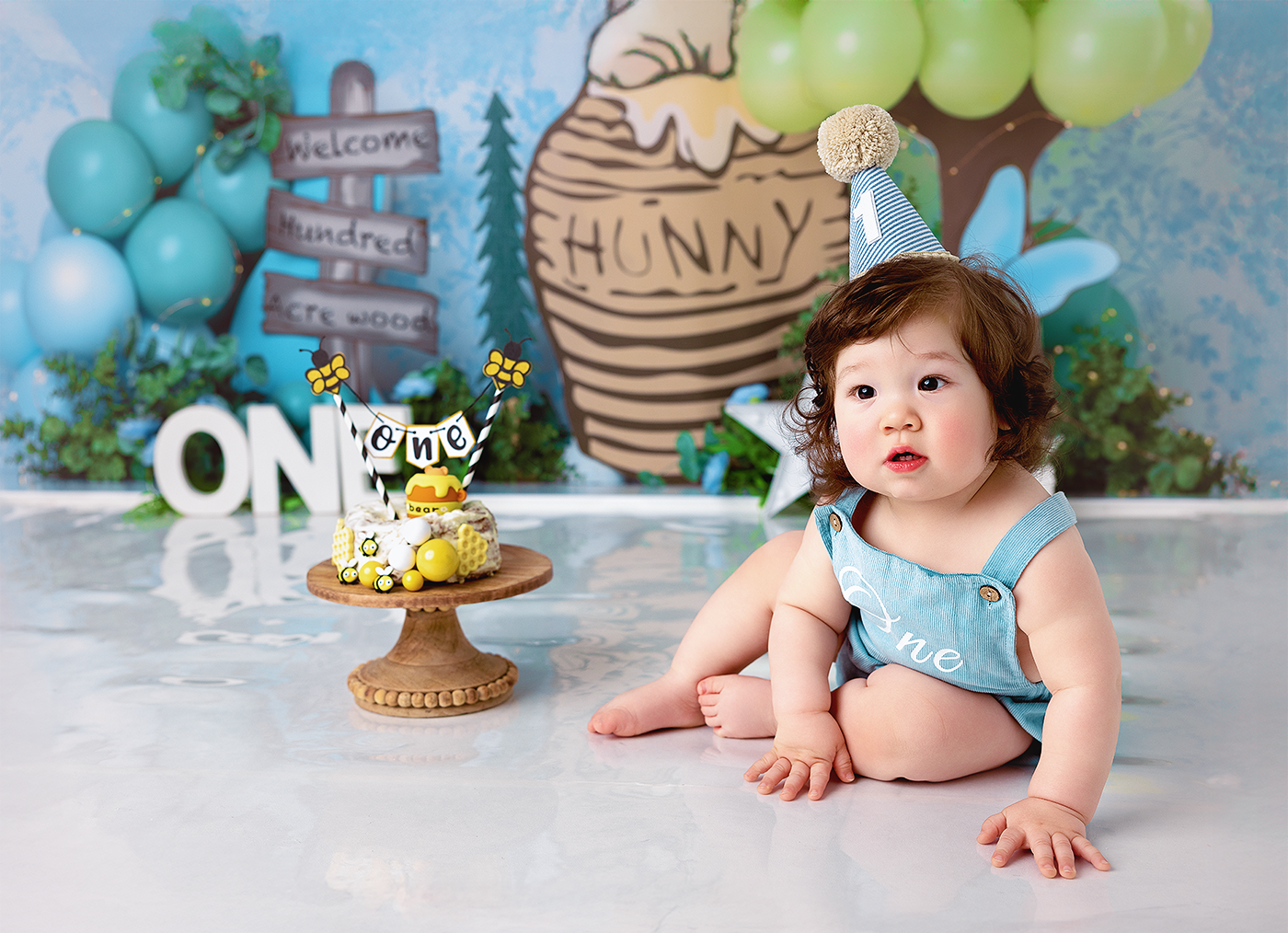 Baby in blue romper sits beside a Winnie the Pooh themed smash cake with balloon decorations for a first birthday.