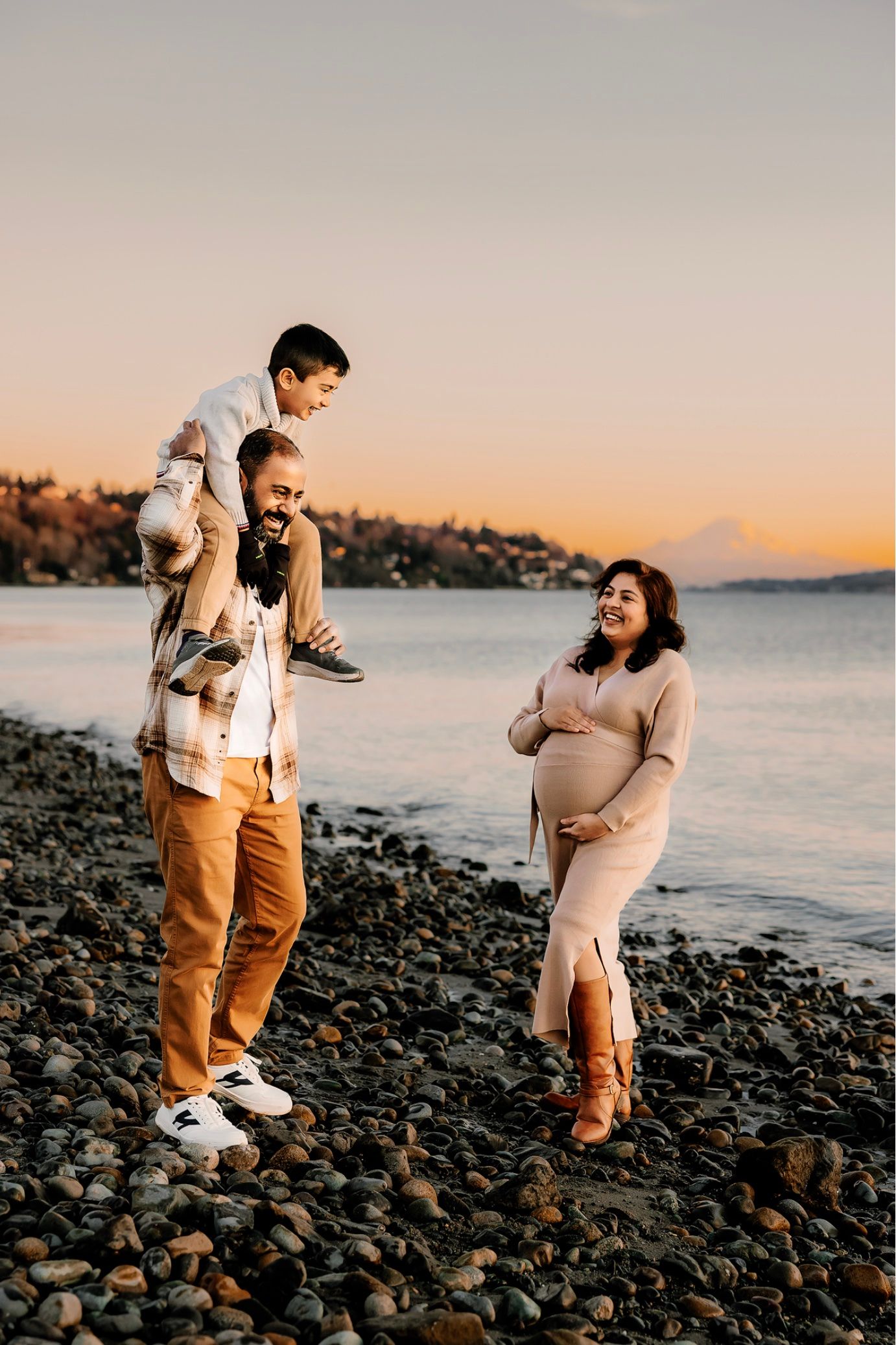 Pregnant woman and man with child on shoulders smile on rocky beach at golden hour sunset.