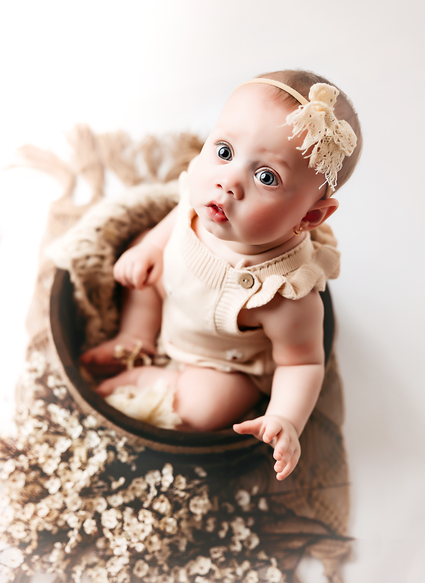 A baby in a cream-colored outfit lies in a wooden bowl surrounded by delicate white floral blossoms.