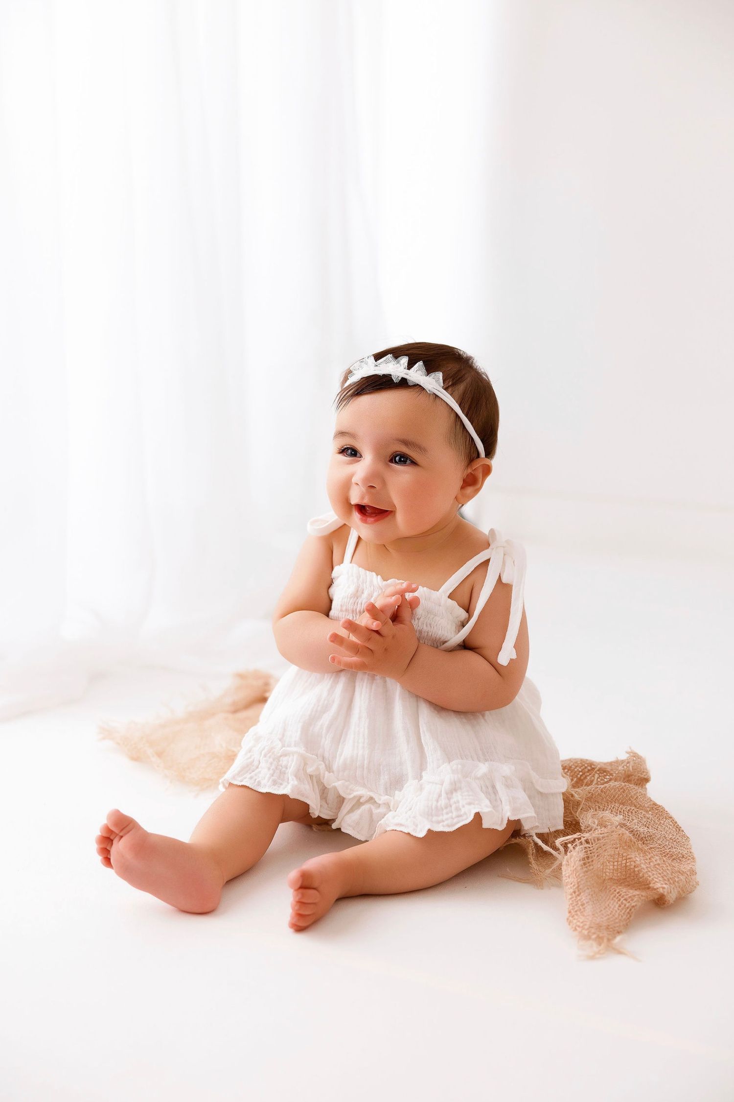 A baby in a white lace dress and headband sits and smiles on a cream-colored backdrop during a photo session.