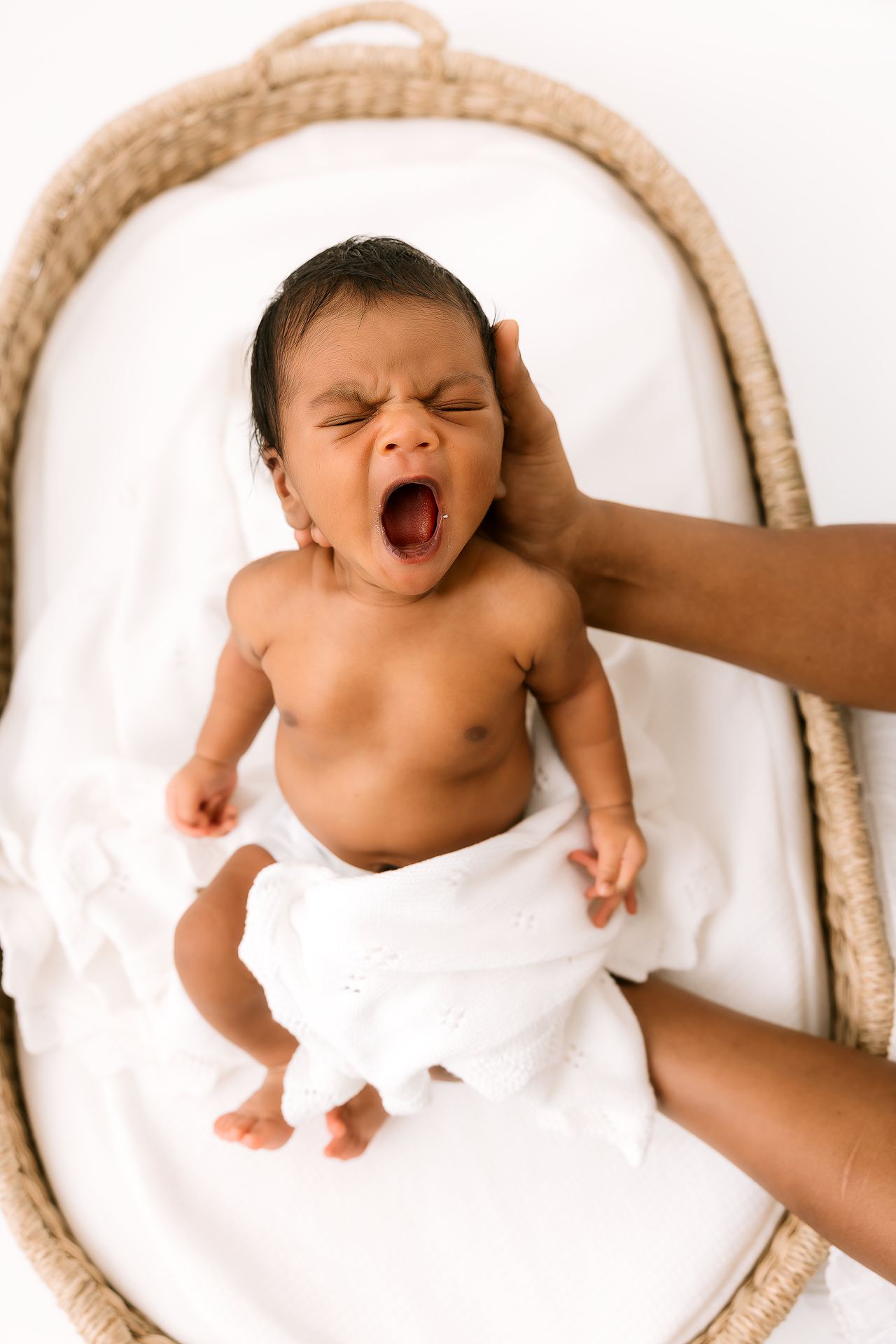 Newborn baby yawning while lying on white blanket with parent's hand visible Everett studio.