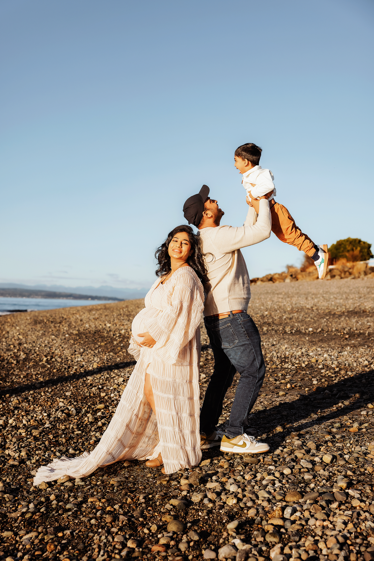 Pregnant woman in white dress poses with partner lifting toddler on rocky beach at golden hour.