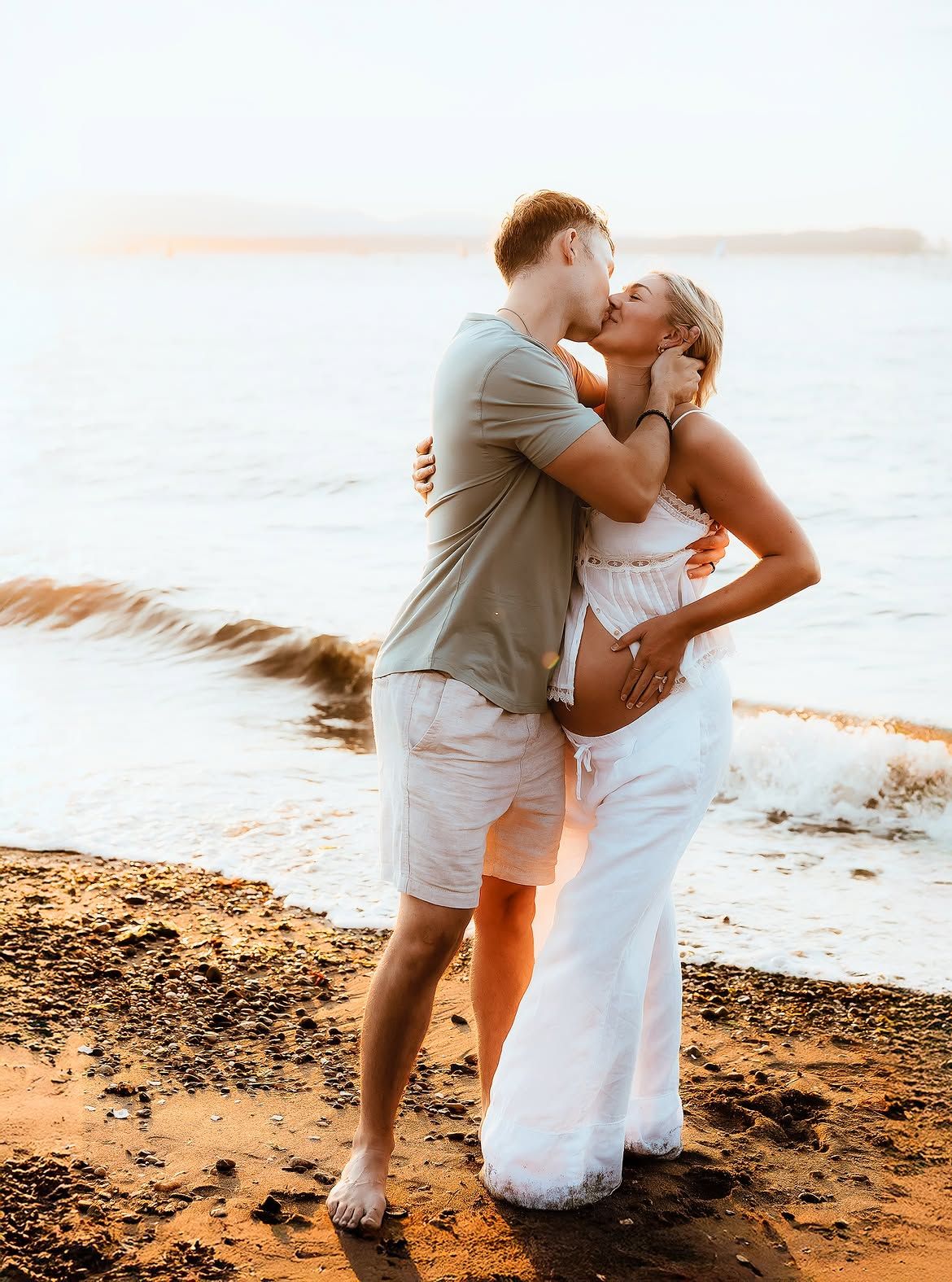 Couple sharing a kiss on a sandy beach at sunset, woman in white dress cradling baby bump.