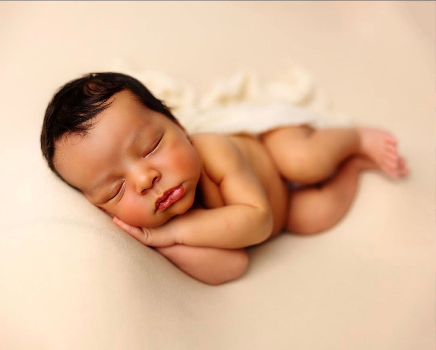 A sleeping newborn baby lies peacefully on soft beige fabric in a minimalist portrait setting.