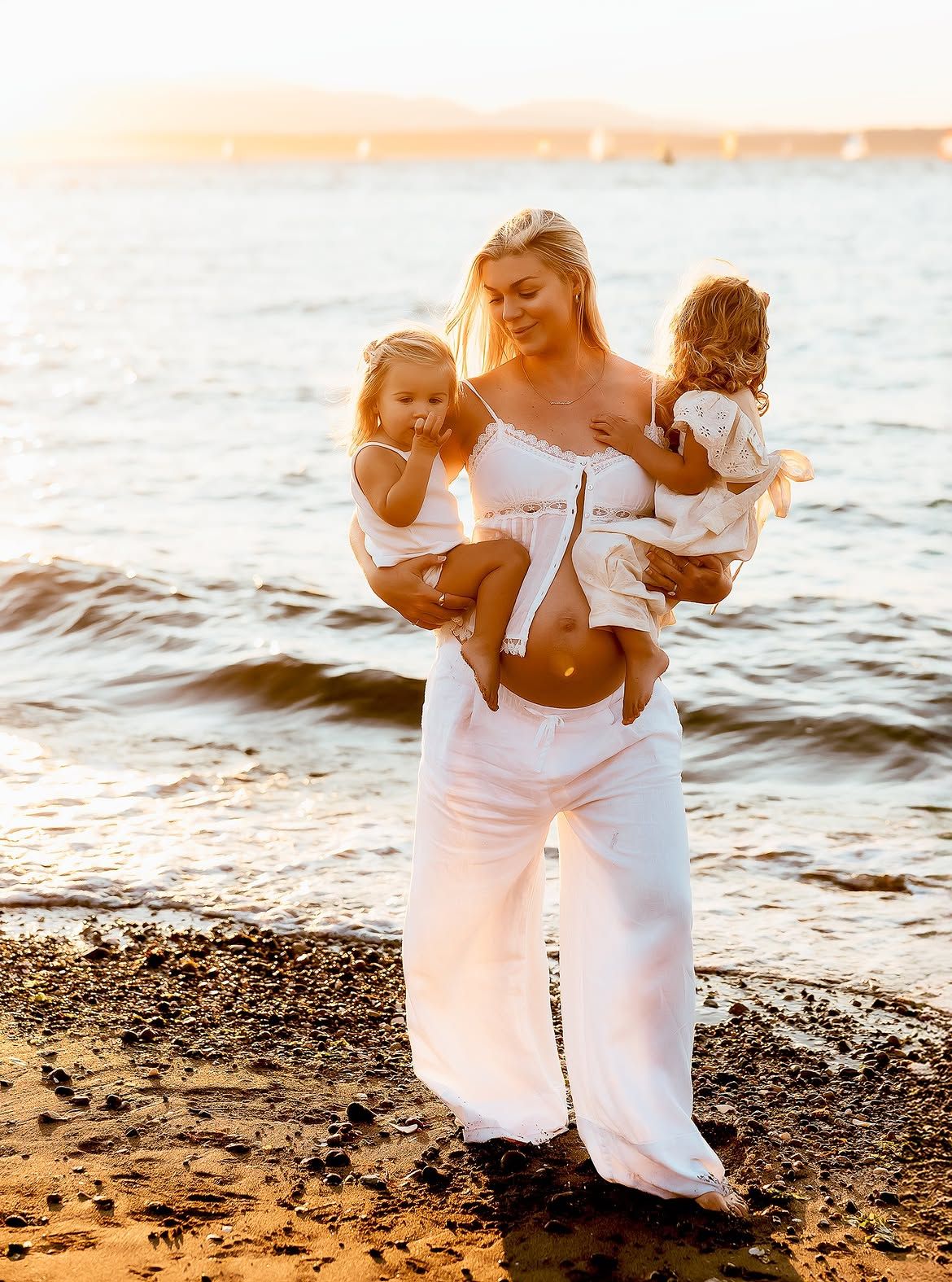 Pregnant woman in white outfit holding two toddlers on a sunny beach at golden hour.