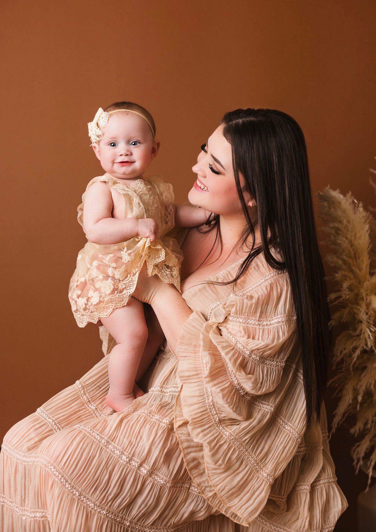 A mother in a beige dress holds her smiling baby girl in a matching outfit during a professional studio portrait session.