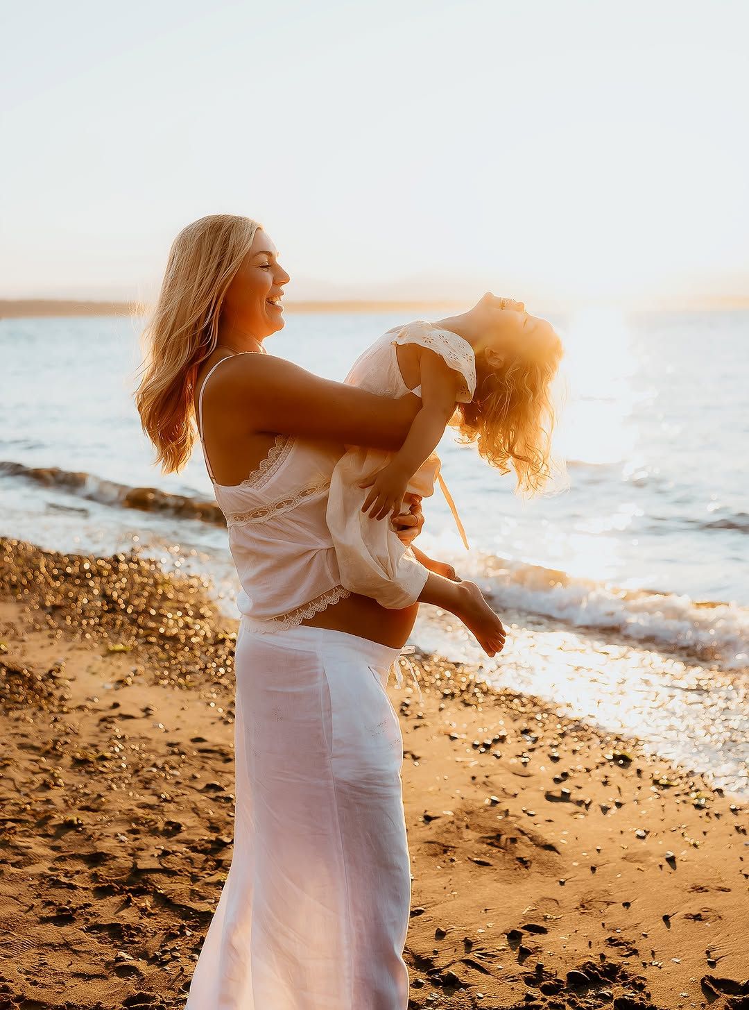 A joyful woman in white lifts a child on a sunny beach, both laughing with golden sunlight and ocean waves behind them.