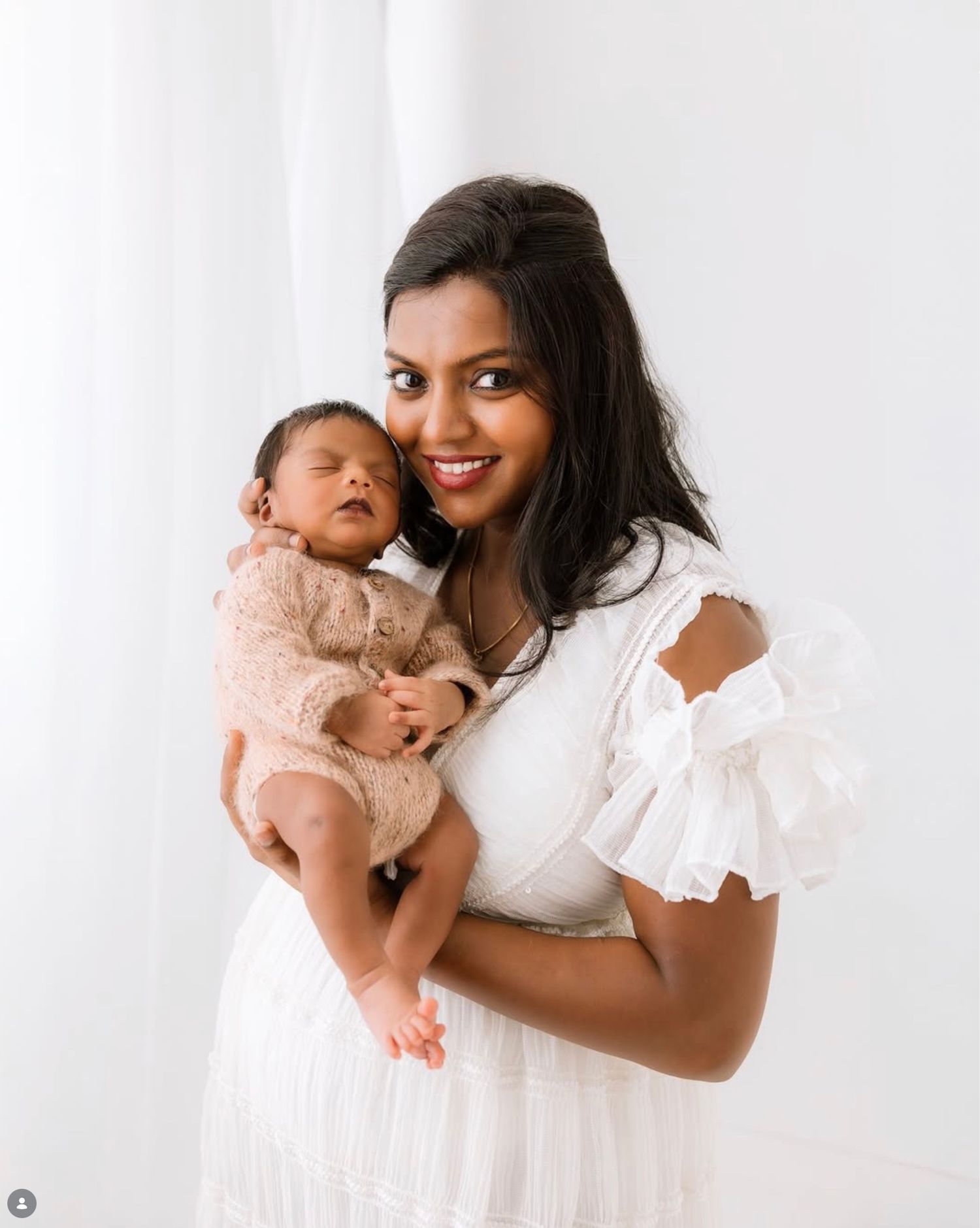 A mother and baby in matching white outfits pose together against a light background for a tender portrait.