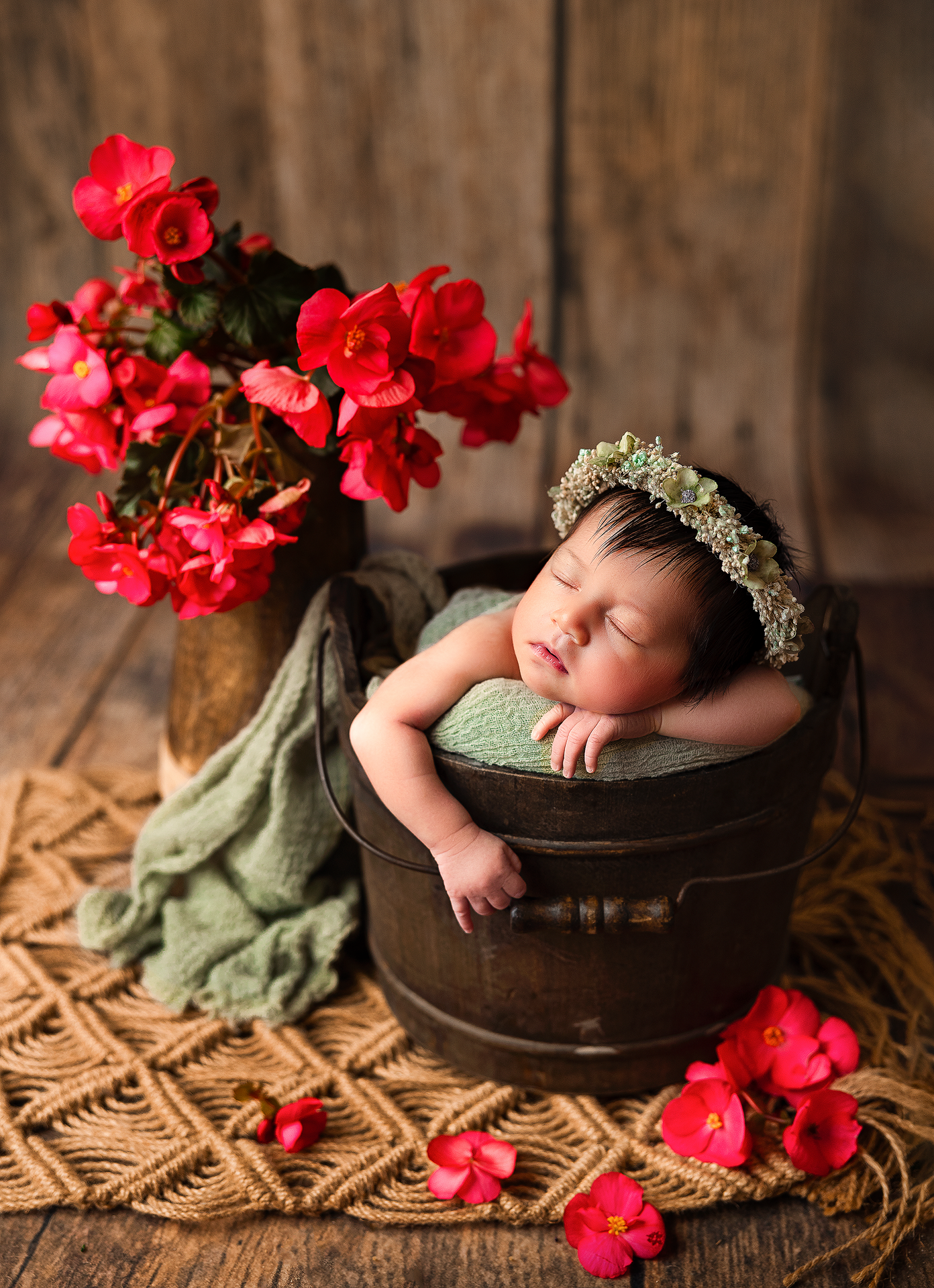 A sleeping baby in a wooden bowl surrounded by vibrant red flowers on a textured beige backdrop.