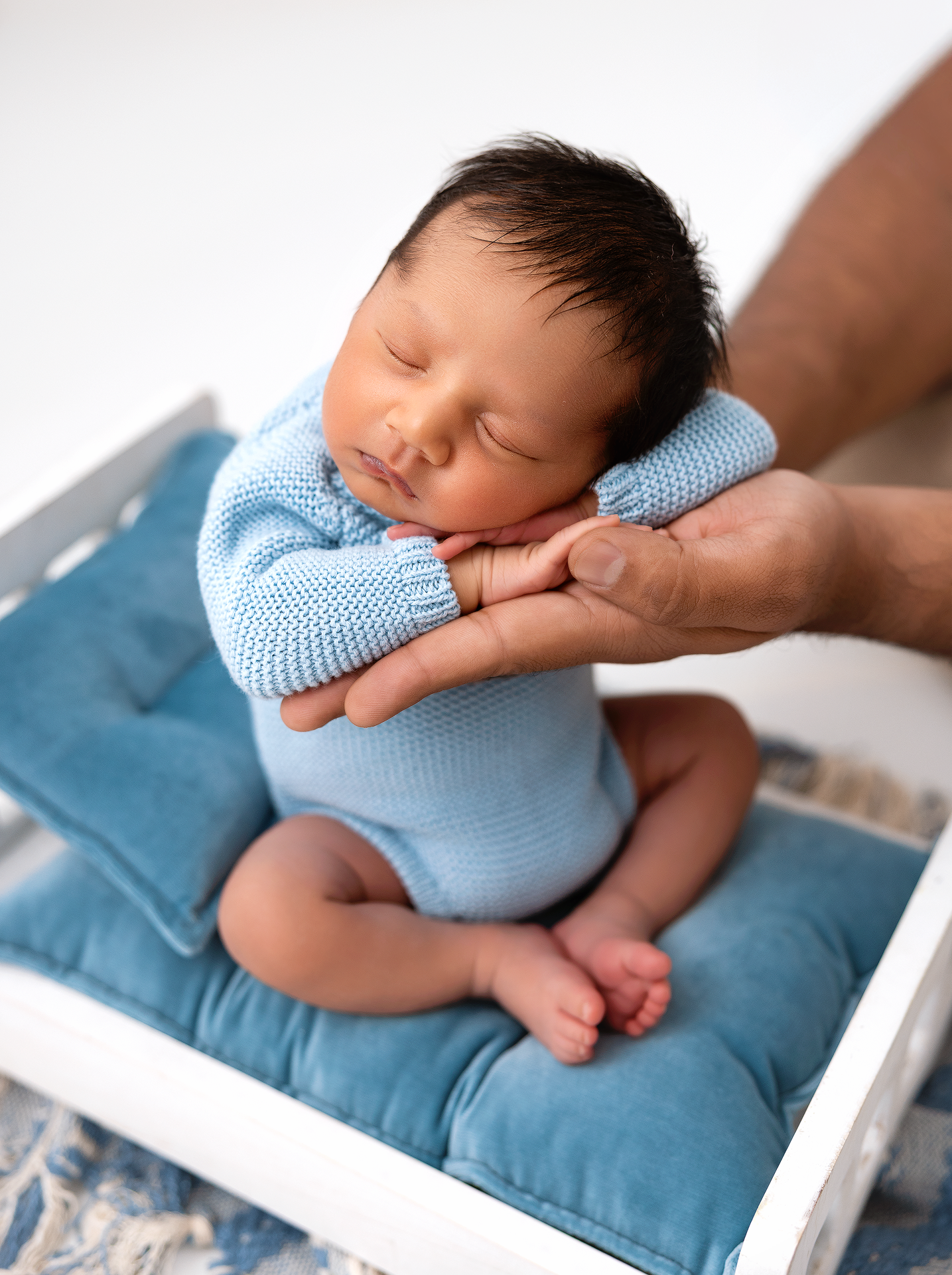 Sleeping newborn baby wearing light blue sweater rests peacefully on a teal cushion during professional photo session.