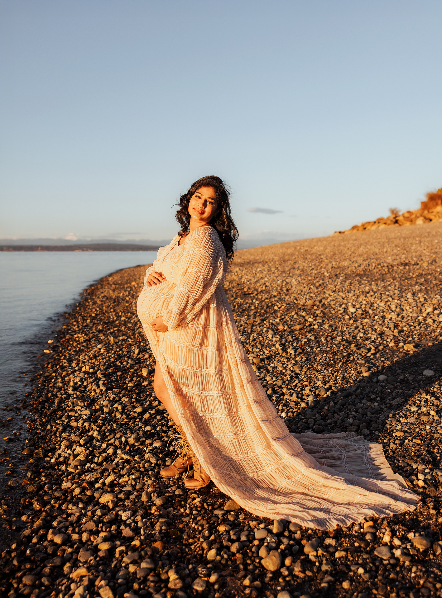 Pregnant woman in flowing pink dress cradling belly on rocky beach at golden hour sunset.