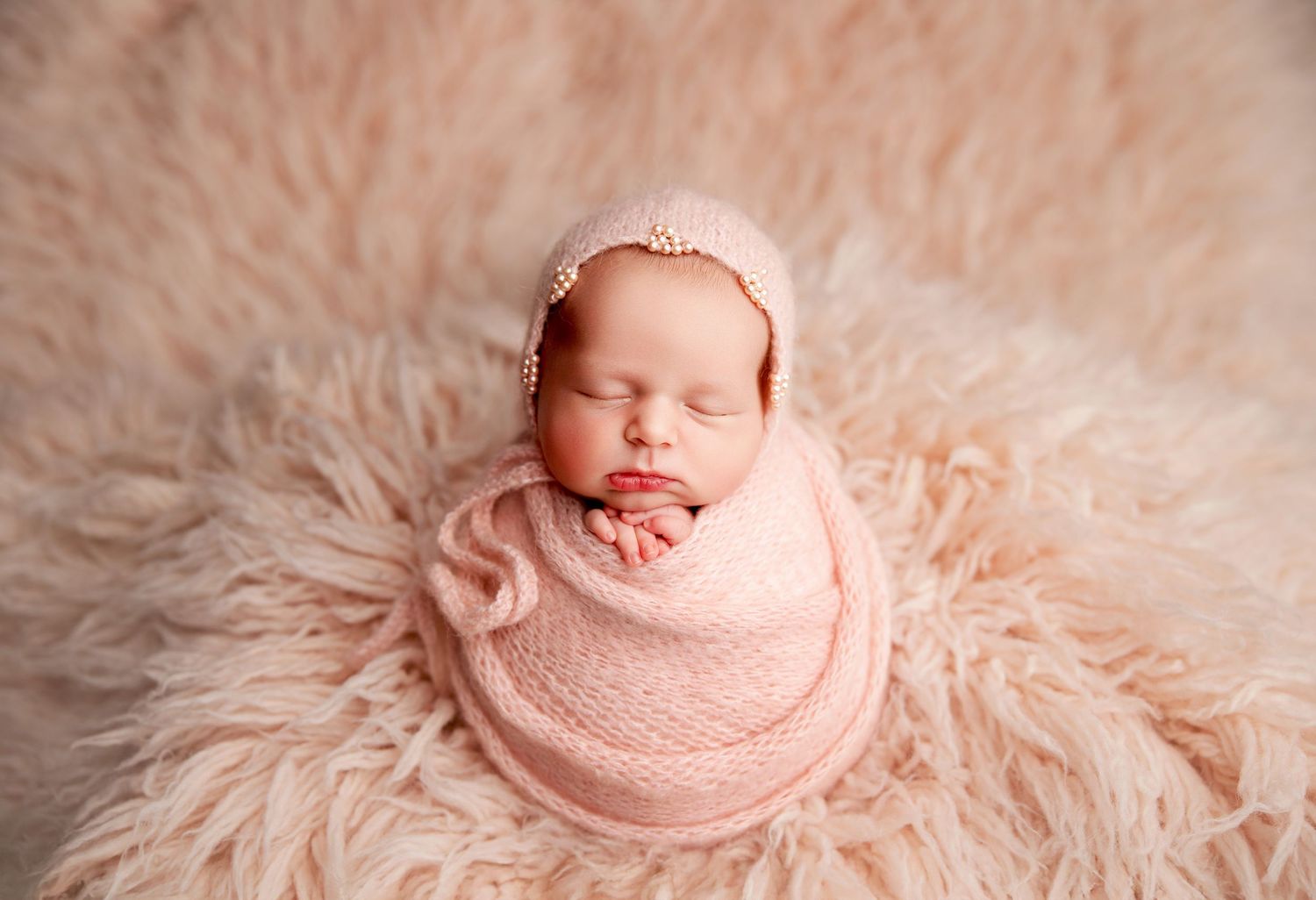 Sleeping newborn baby wrapped in pink knit swaddle with floral bonnet on fluffy pink fur blanket.