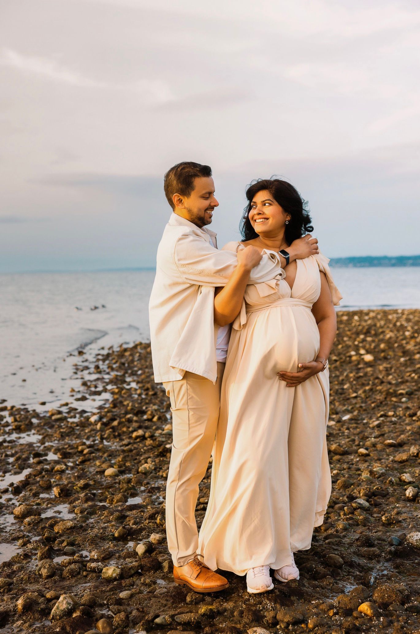 Couple in matching cream outfits posing on rocky beach, pregnant woman cradling bump at golden hour.