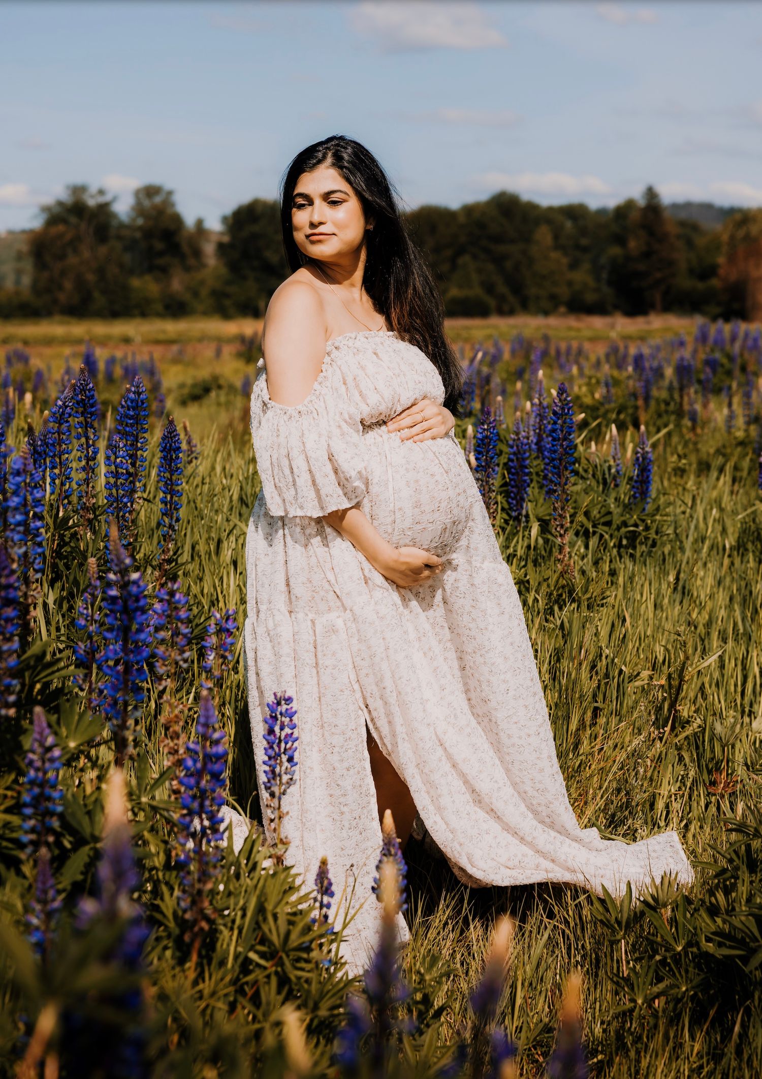 A person in a flowing white dress poses gracefully among vibrant purple lupine flowers in a sunlit meadow field.