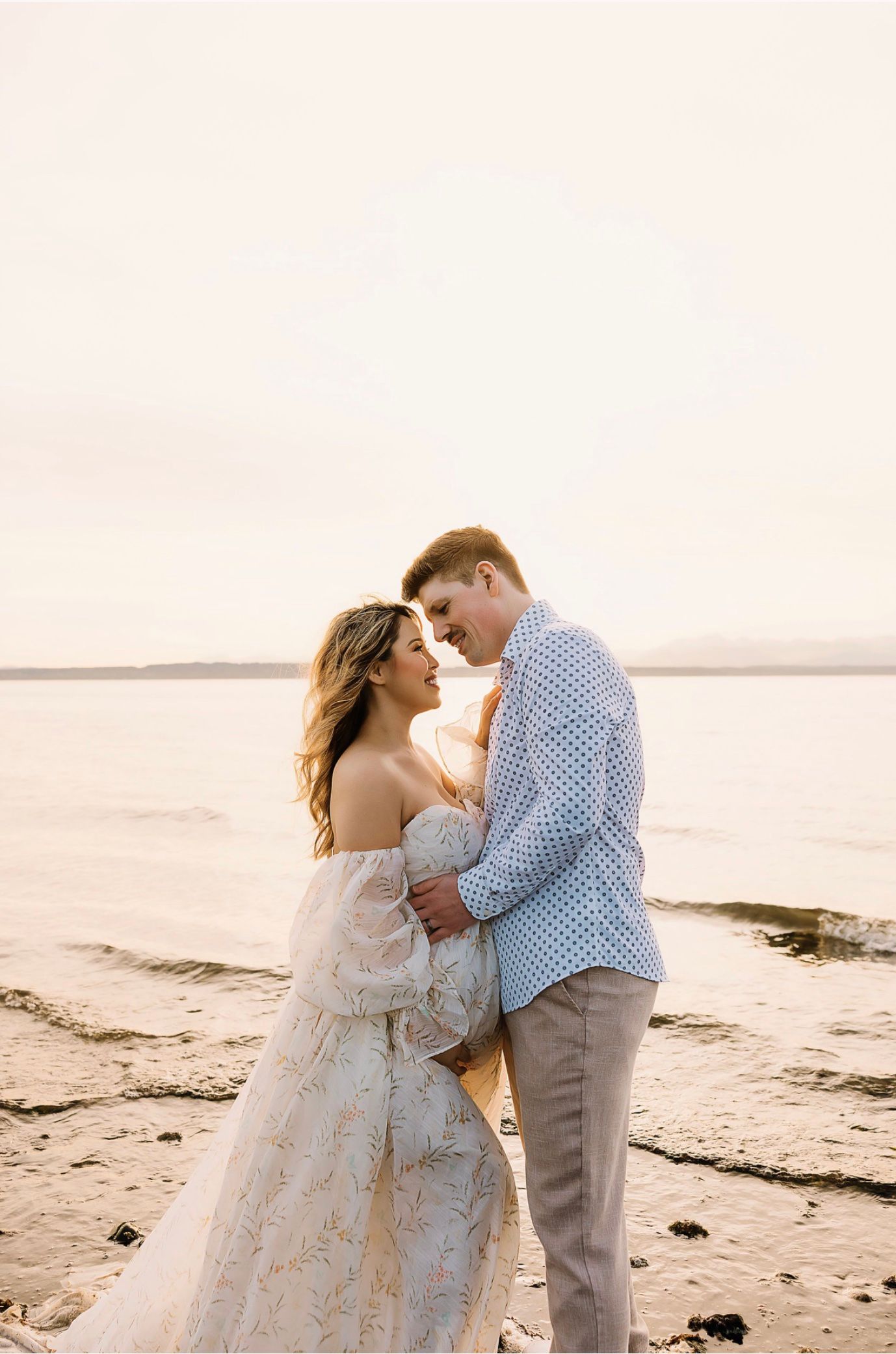 Couple embracing on a sandy beach at golden hour, woman in floral gown, man in blue shirt and gray pants.