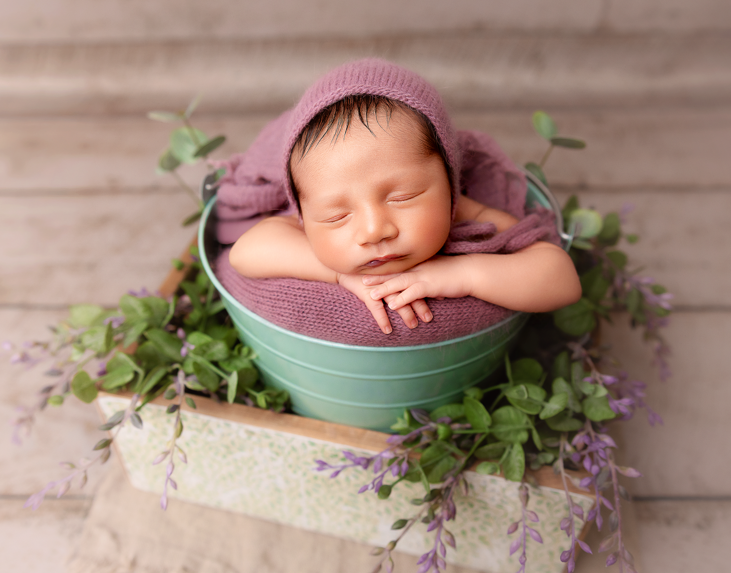 Sleeping newborn baby in teal bucket with purple bonnet, surrounded by green and lavender flowers on wooden backdrop.